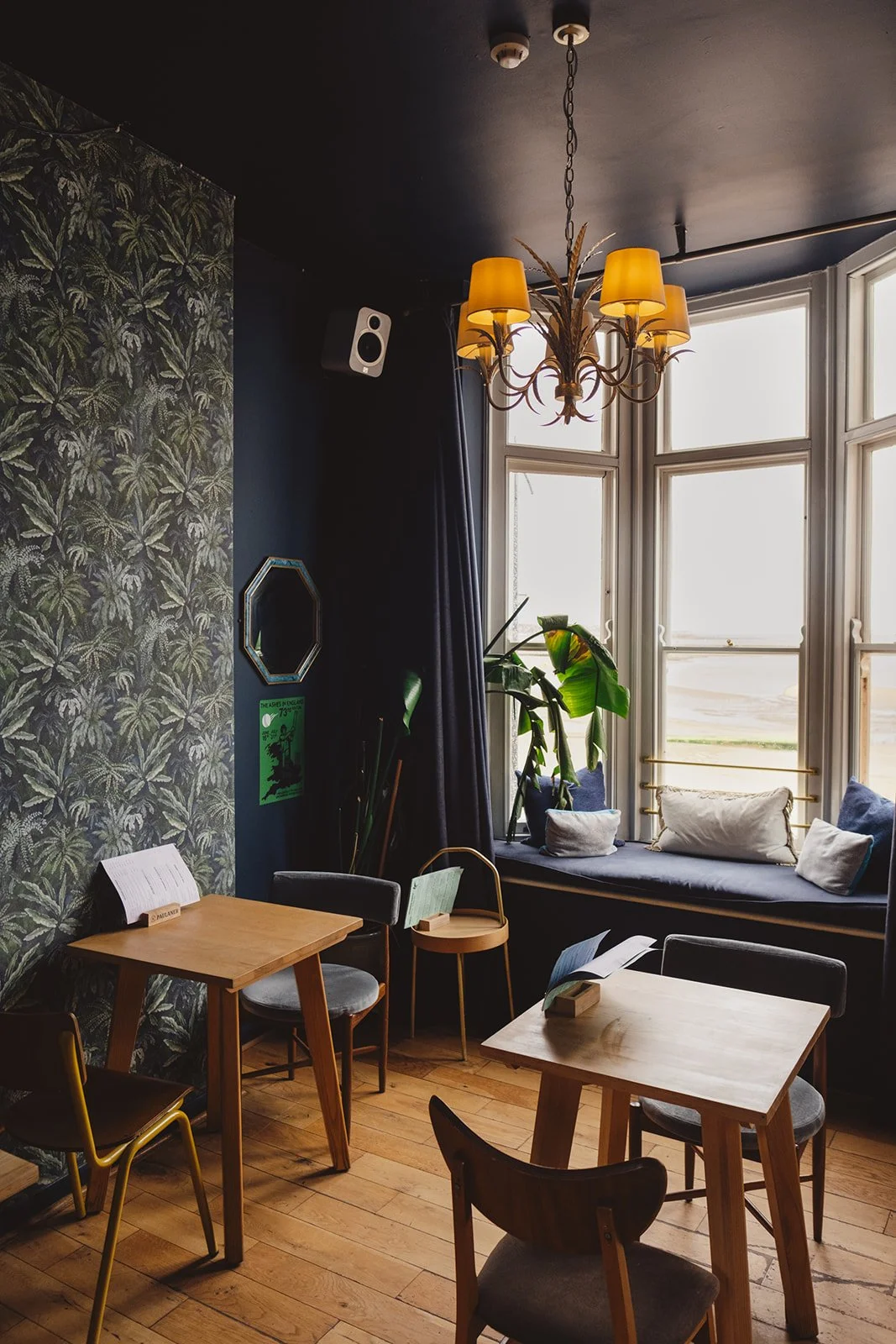 Interior of a cozy cafe or sitting area with wooden tables and chairs, a window seat with cushions, a large window, plants, a chandelier with yellow shades, and wallpaper with a leafy palm pattern.