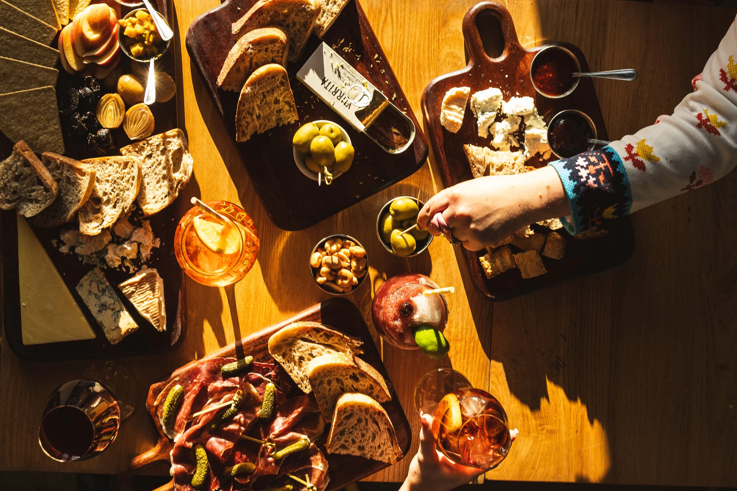 A wooden table filled with various types of bread, cheeses & meats to enjoy as part of Little Swifts drinking food platters.