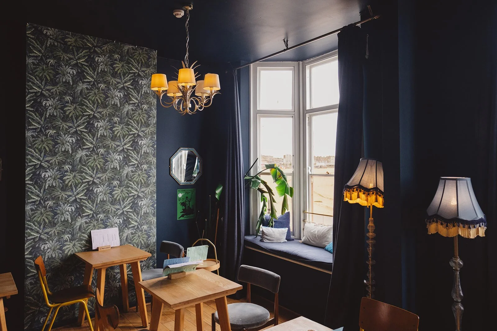 Cozy interior of a cafe with a dark blue wall, patterned wallpaper, a window seat with cushions, a hanging chandelier with yellow shades, and two standing lamps with fringed shades, near a bay window with curtains and potted plants.