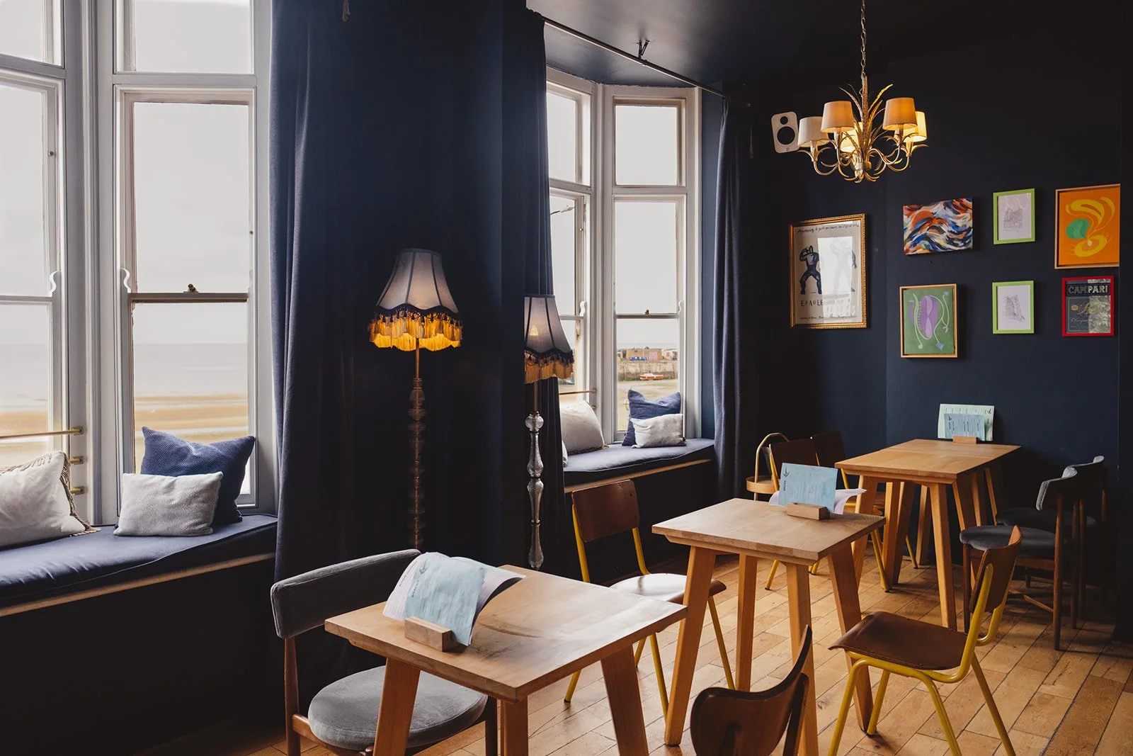 A cozy restaurant interior with wooden tables, mixed chairs, and window seat cushions, decorated with framed art on navy blue walls and illuminated by vintage lamps and a chandelier.