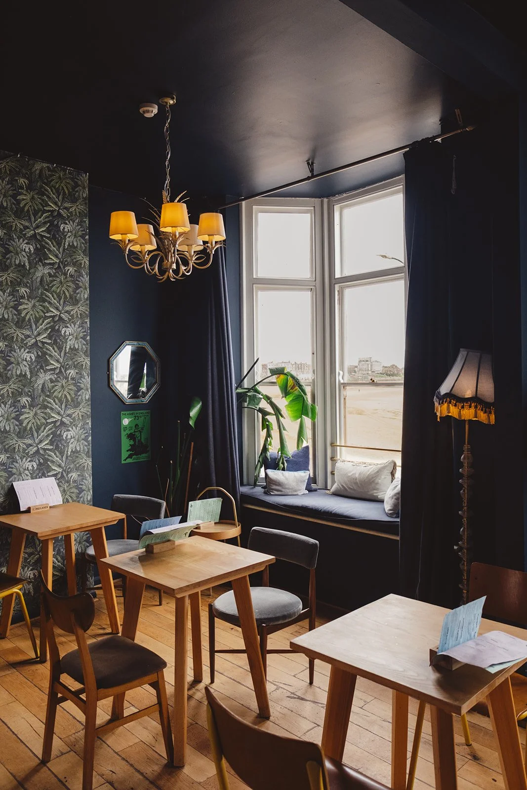 Interior of a cozy bar with wooden tables and chairs, dark walls, a window seat with cushions, plants, a chandelier, a standing lamp, and a window with curtains.