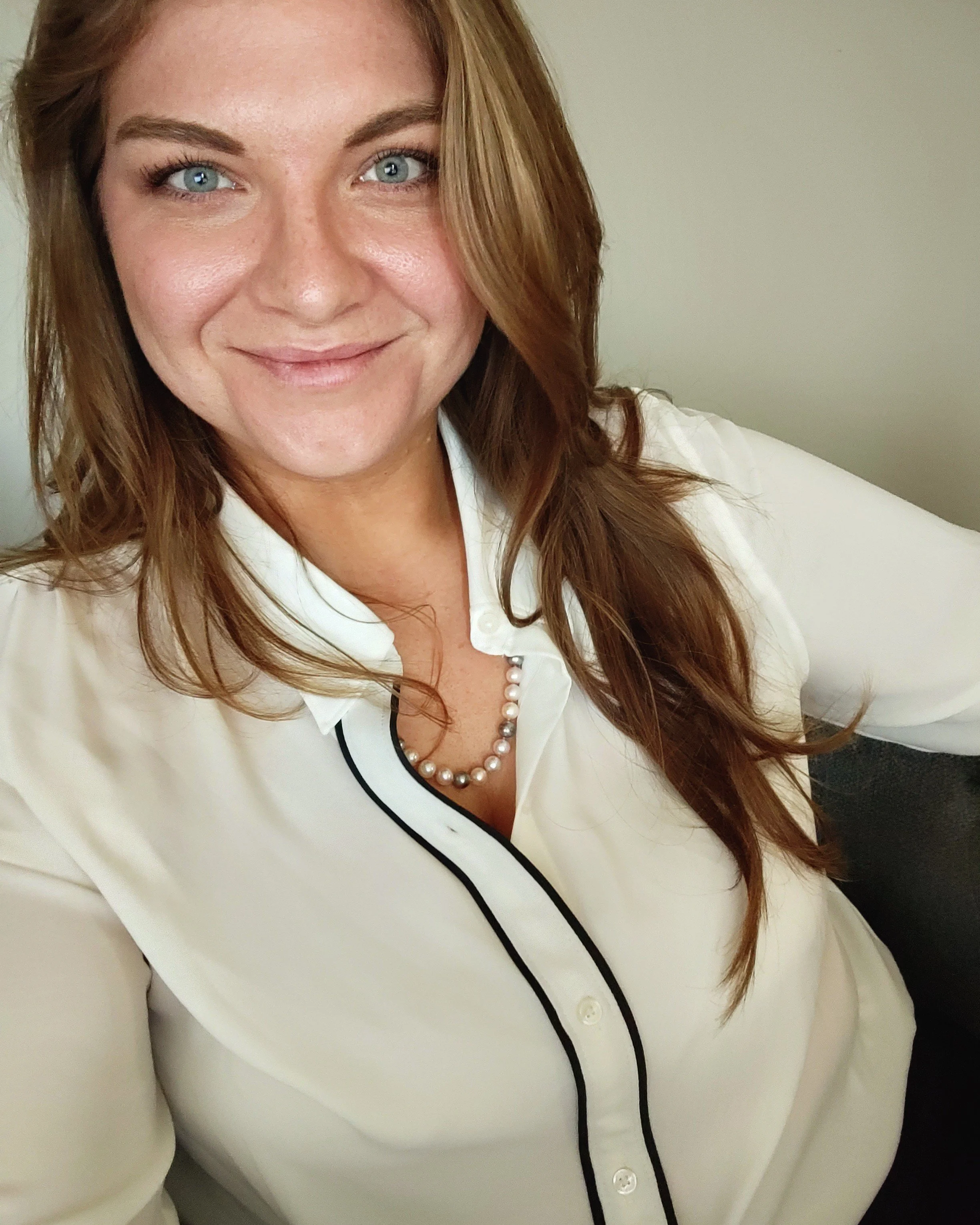 A woman with long, wavy auburn hair and blue eyes smiling at the camera, wearing a white blouse with black piping and a pearl necklace, sitting on a gray sofa against a light-colored wall.