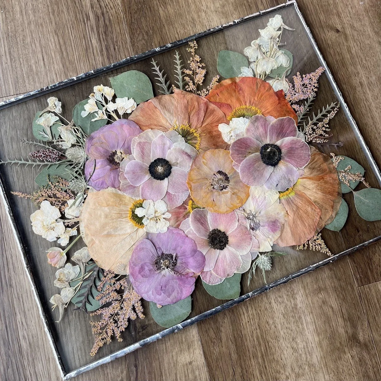 Image is of a bridal flower arrangement, pressed and preserved between two panes of glass. It is soldered and sealed with silver metal solder, and includes a chain for hanging. The flowers include pressed poppies, anemone, ranunculus, and stock.
