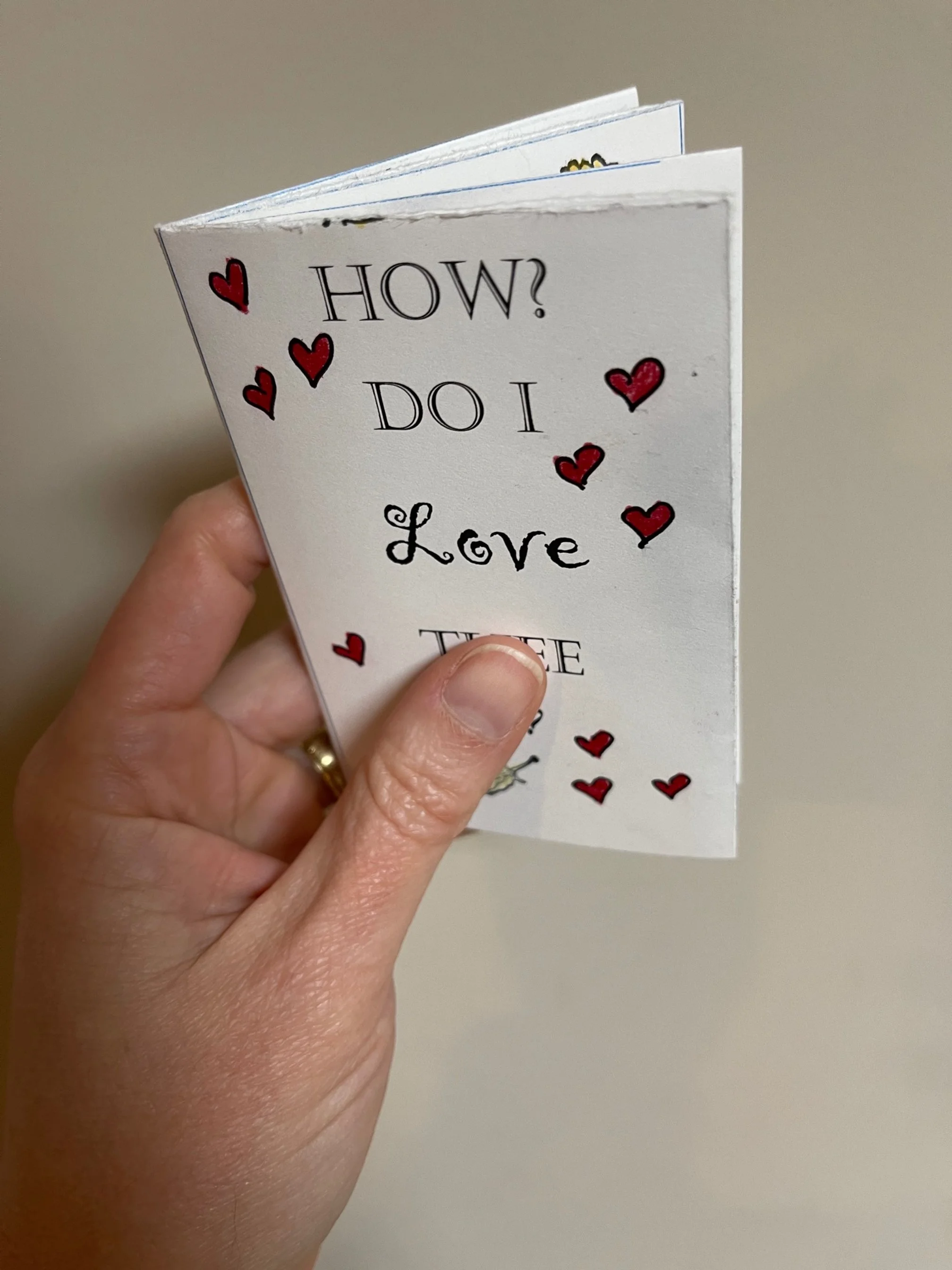 A hand holding a small folded zine. The front page of the zine reads typed, "How do I love thee?" with drawn red hearts.