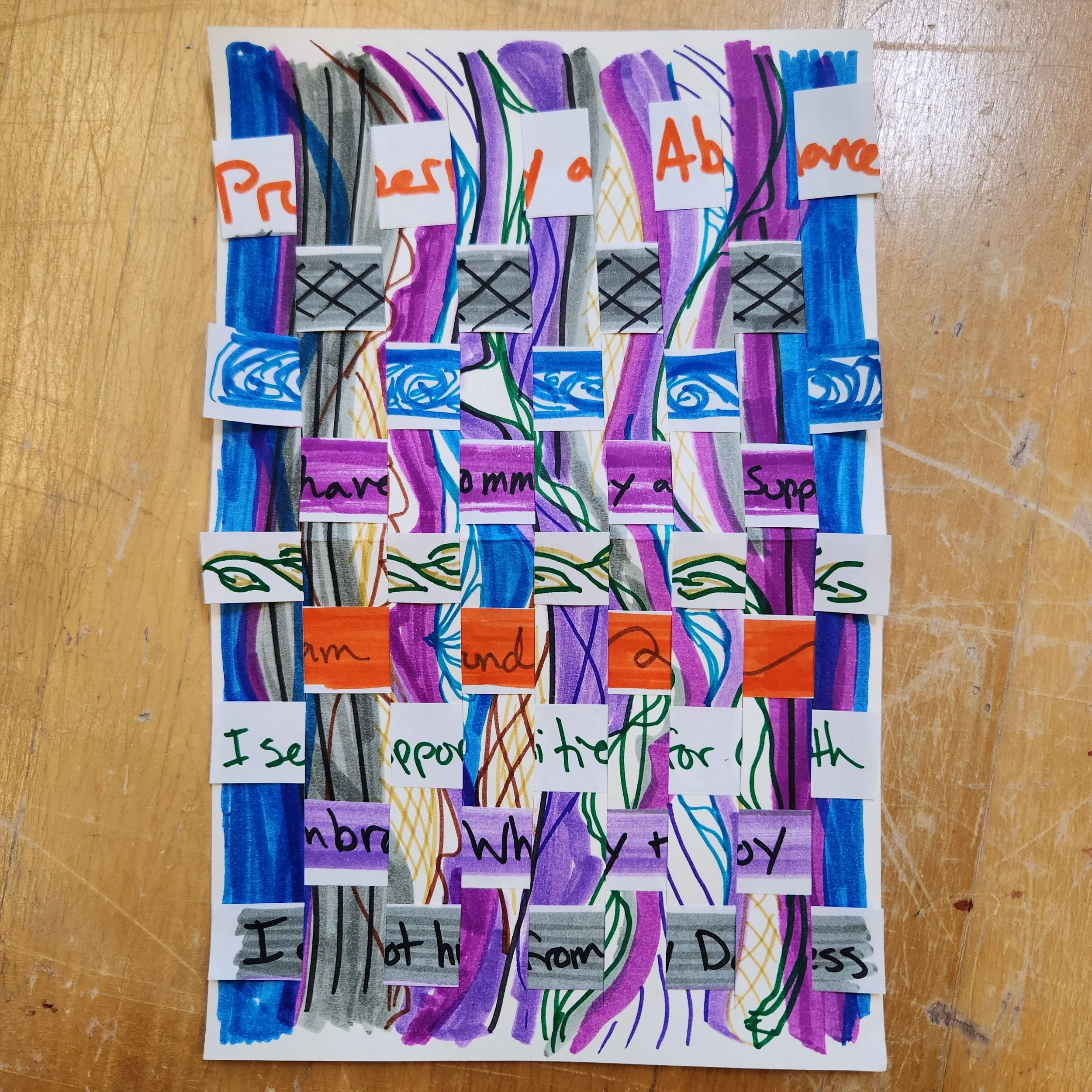 Vertical and horizantal strips of white paper weaved together, with purple, blue, orange, and green marker designs drawn on.