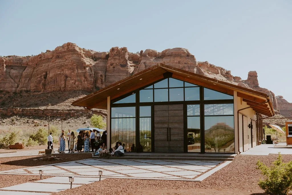 Wedding guests gathered outside the Glass Venue at The Red Earth Venue in Moab, Utah, with dramatic red rock cliffs rising behind the building during a summer wedding.