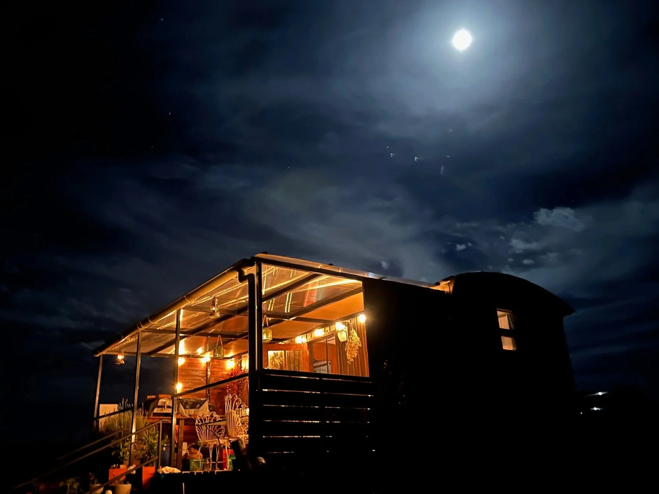A house with warm interior lights on a dark night, under a partly cloudy sky with the moon overhead.
