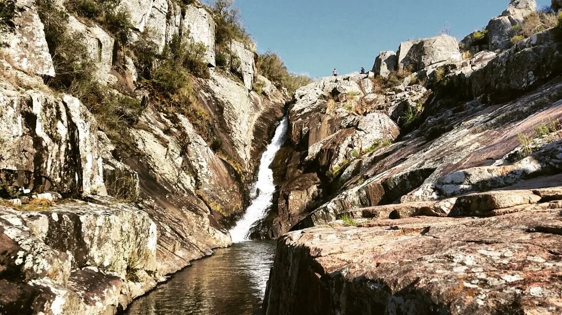 A waterfall flowing down rocky terrain into a small pool of water surrounded by large rocks and boulders.