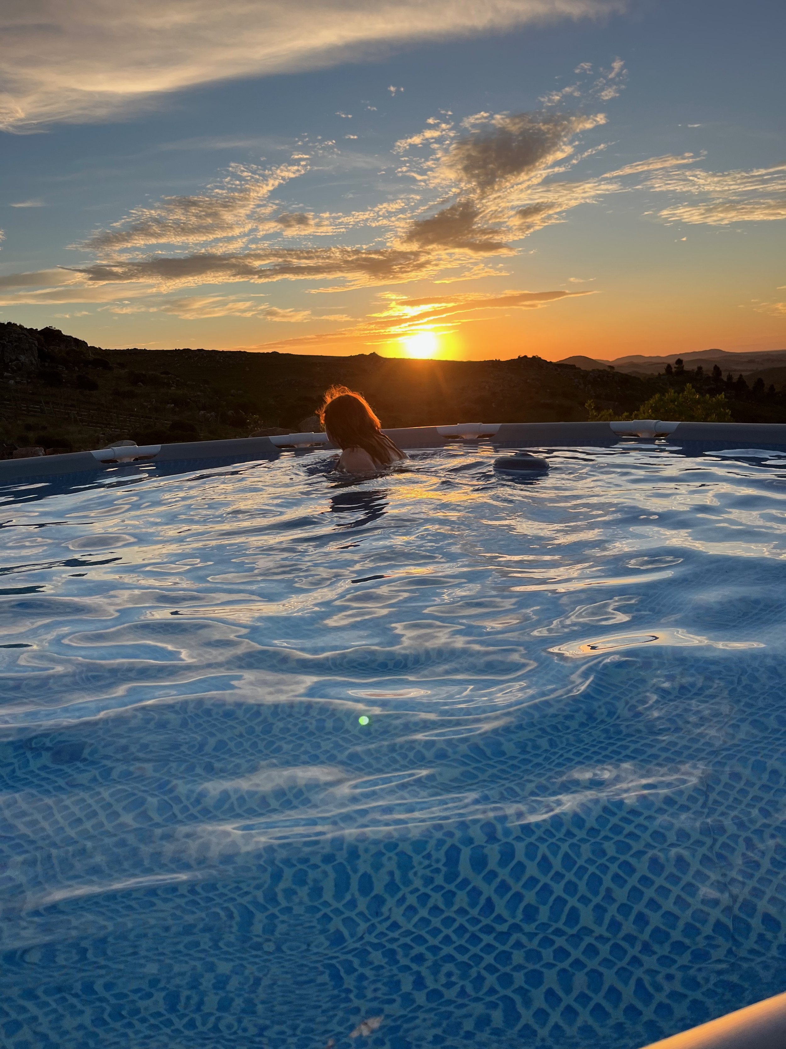 Person swimming in a pool during sunset, with hills and a partly cloudy sky in the background.