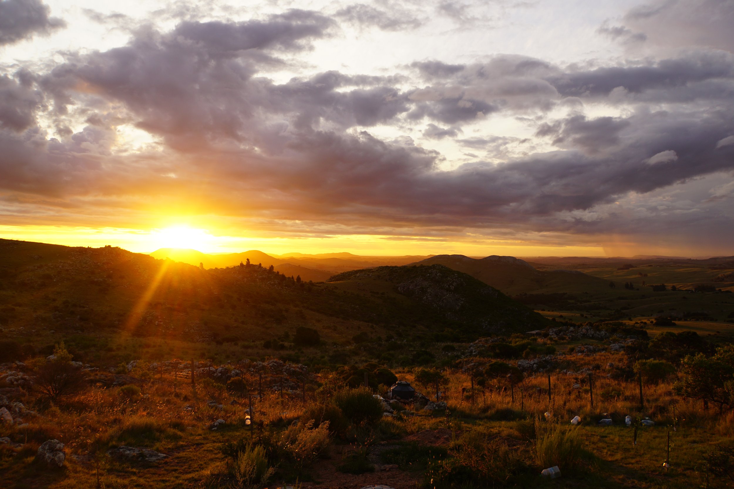 Sunset over rolling hills with partly cloudy sky and distant mountains.