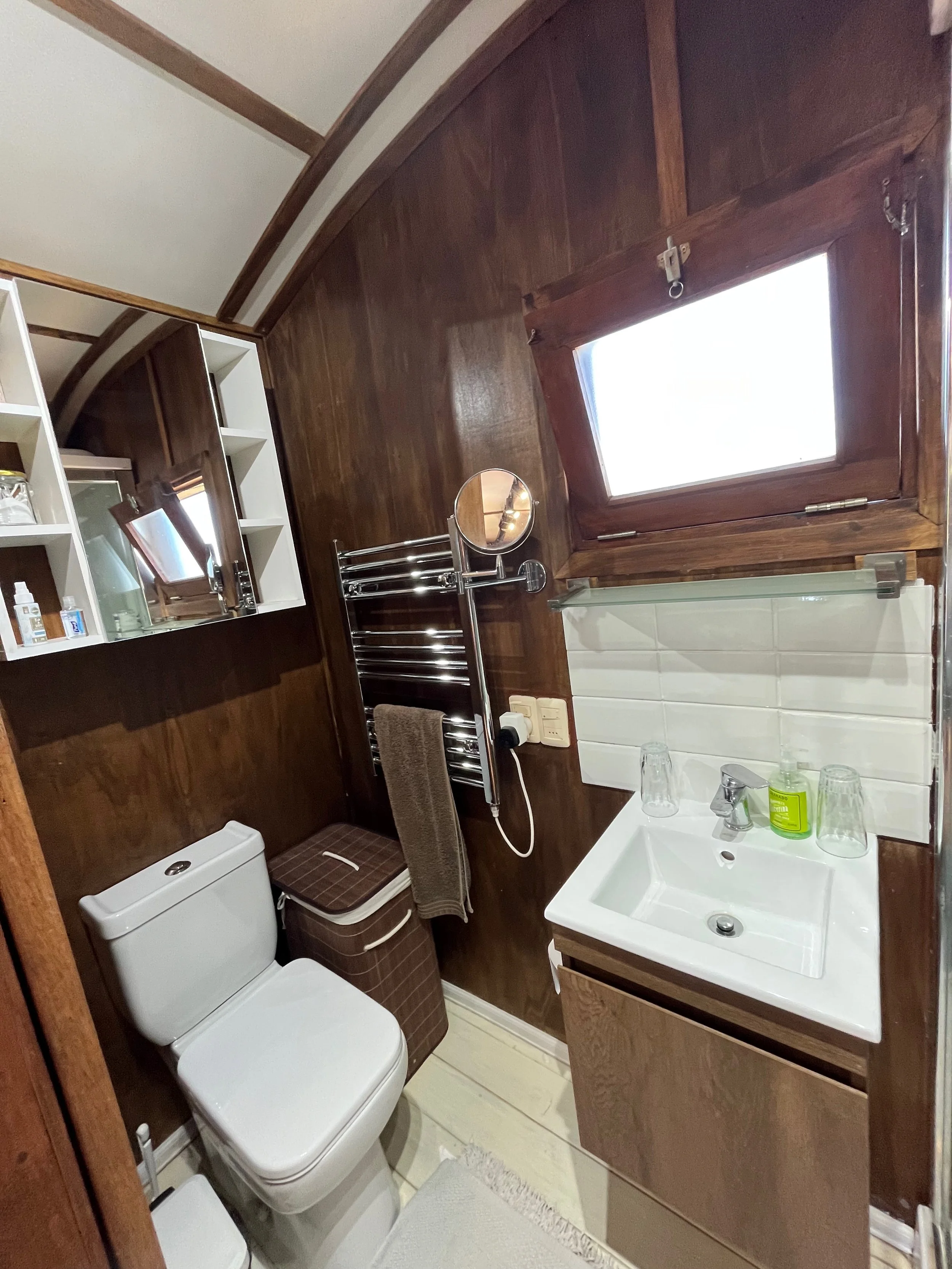 Small bathroom with wooden walls and ceiling, featuring a white toilet, a small white sink with a mirror, and a window with wooden framing. There is a towel rack with a brown towel, a small trash bin, and some toiletries on a shelf.
