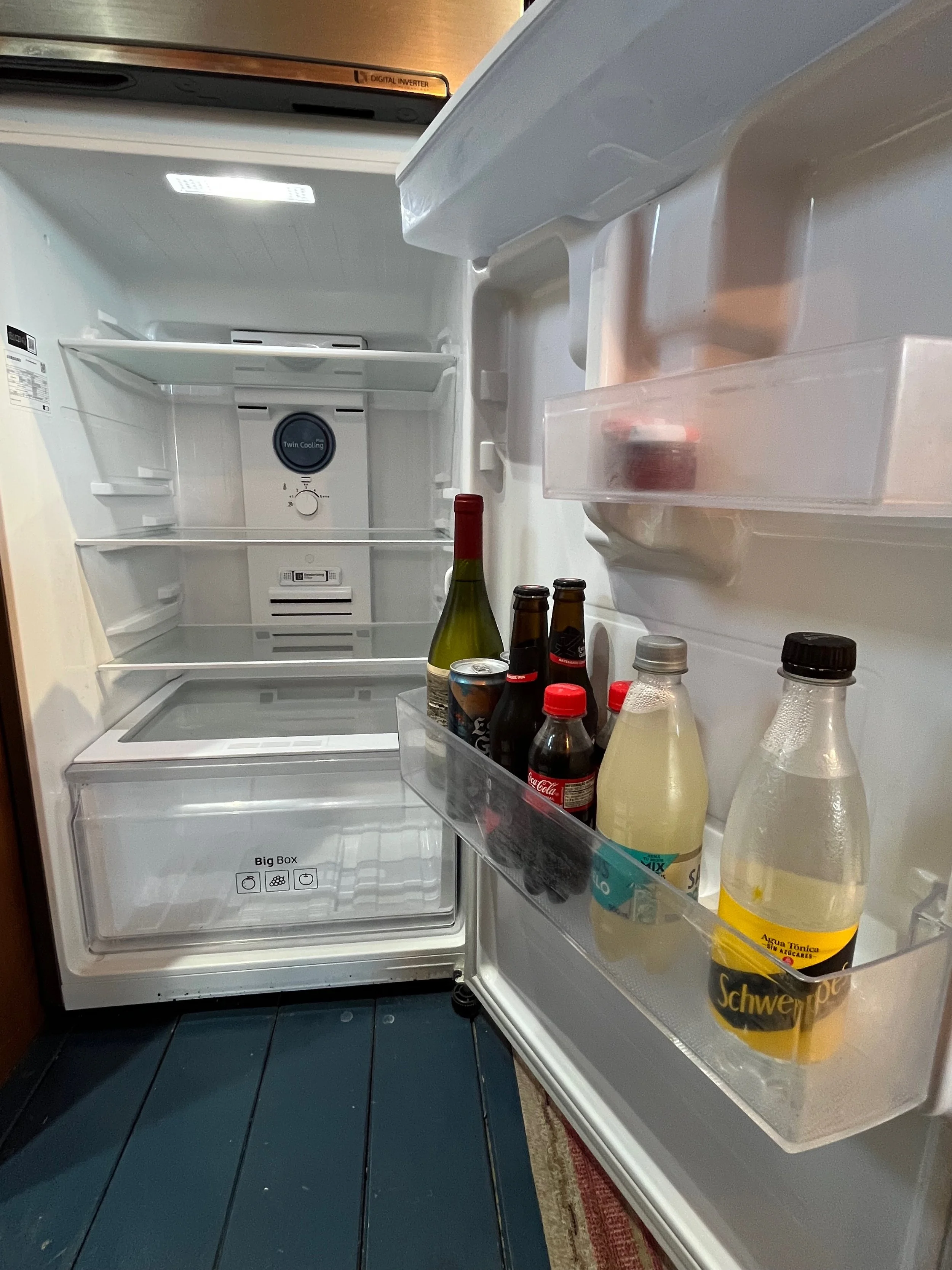 Open empty refrigerator with some bottled beverages on the door shelf, including soda and tonic water, with a wine bottle inside.