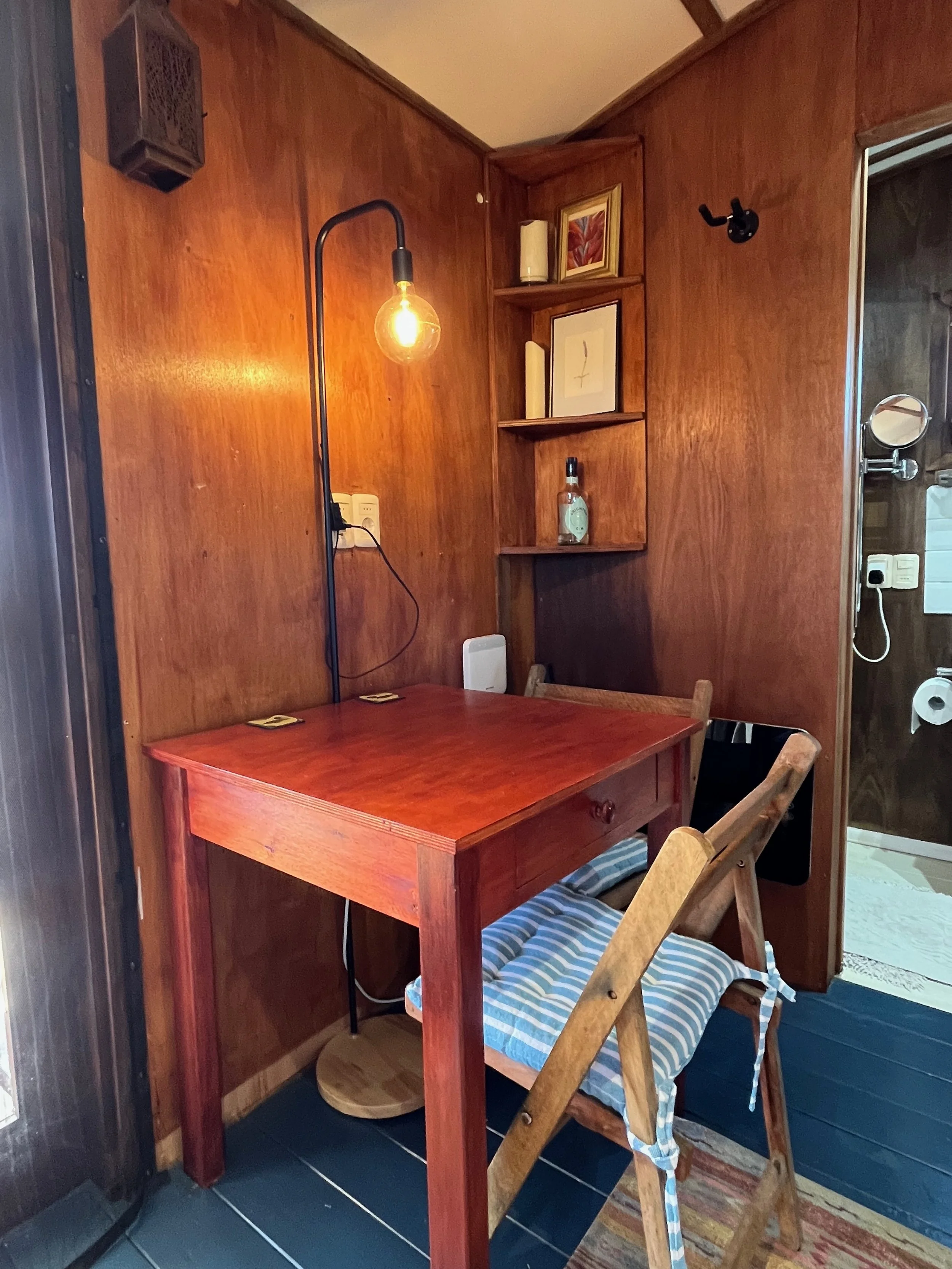 A small wooden desk with a matching chair and a striped cushion. A black floor lamp with an exposed filament bulb is positioned over the desk. The wall behind is made of wood paneling and has built-in shelves with decorative items and a framed pictur