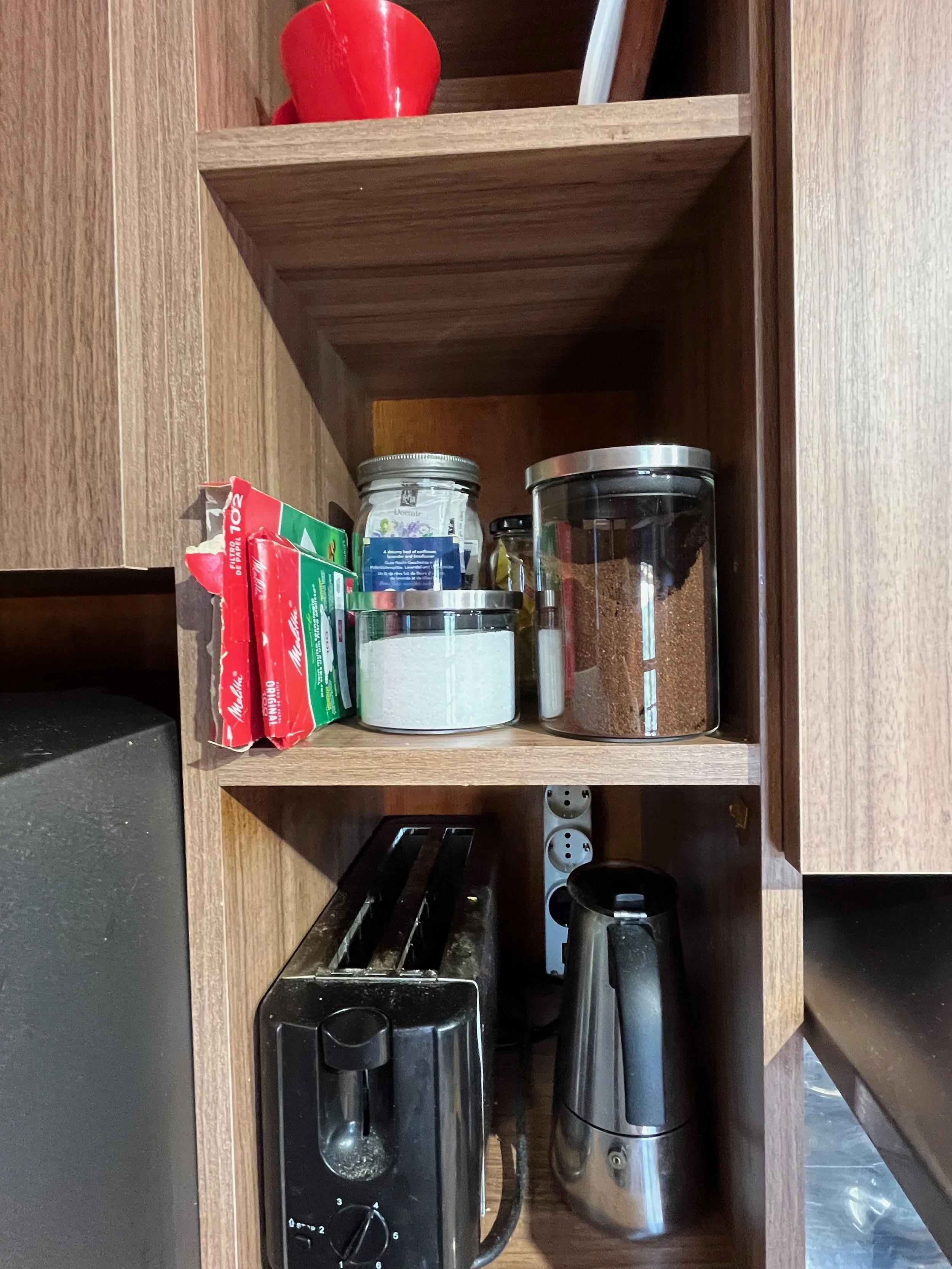 Kitchen cabinet with coffee maker, toaster, and jars of spices and coffee, above a power strip with multiple outlets.