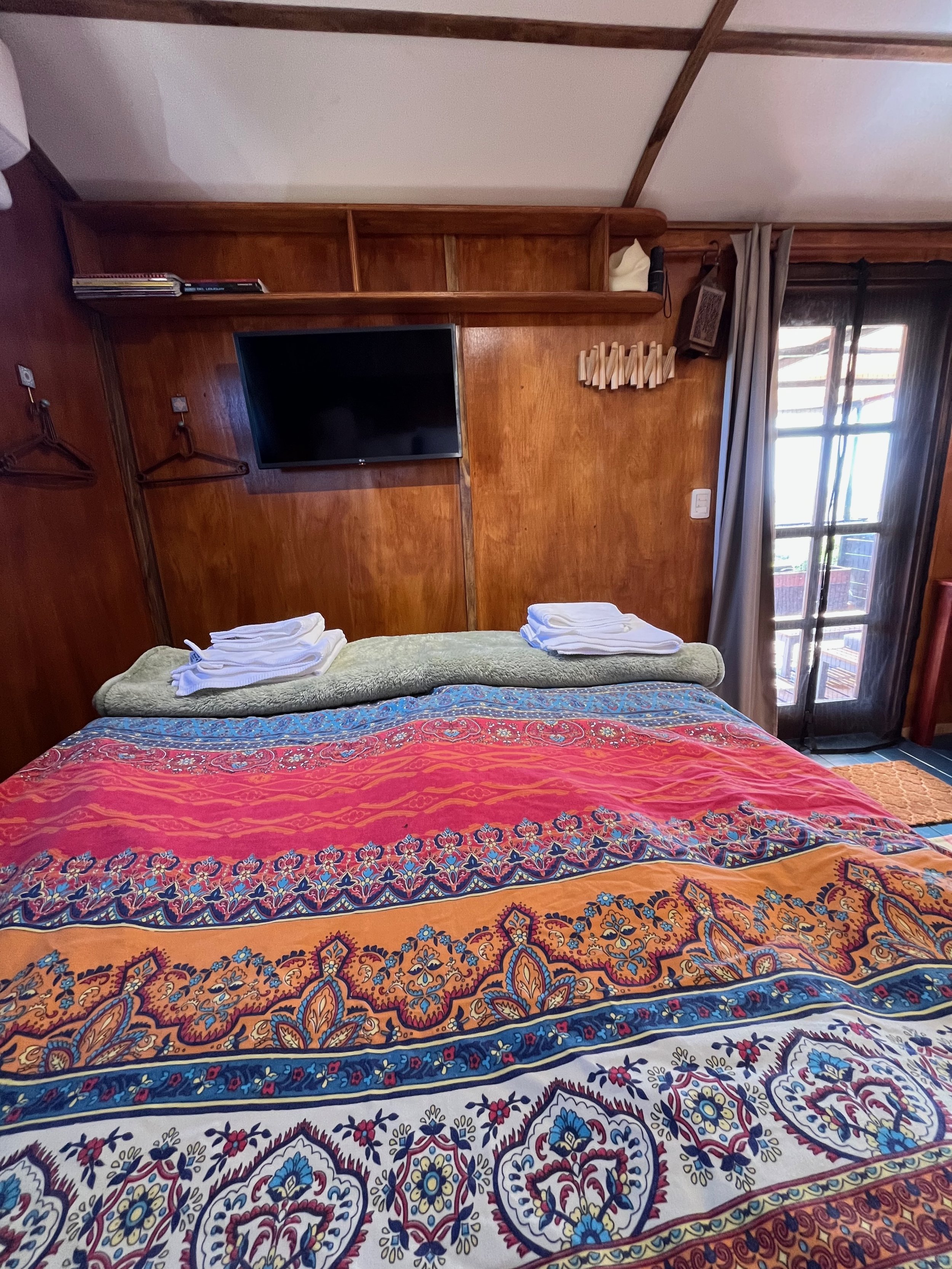 A cozy hotel room with a bed covered by a colorful patterned quilt, set against a wooden wall with a mounted TV and shelves. There are folded towels on the bed, and a sliding glass door with curtains leading to a balcony.