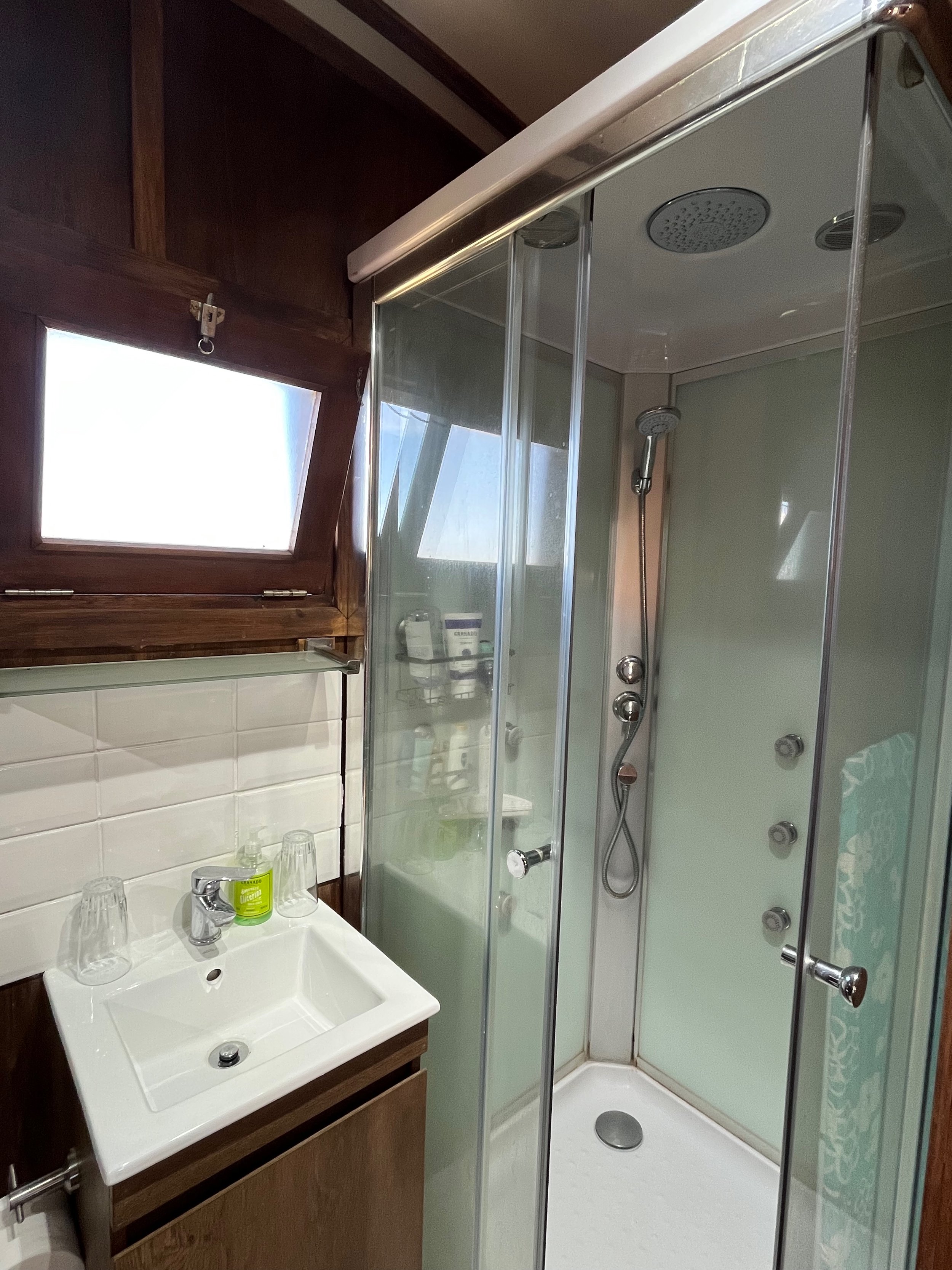 Small bathroom with wooden wall panels, a rectangular white sink with a chrome faucet, two glasses, a green soap bottle, and a mirrored shelf above it. There is a frosted glass corner shower with sliding doors, featuring multiple showerheads and cont