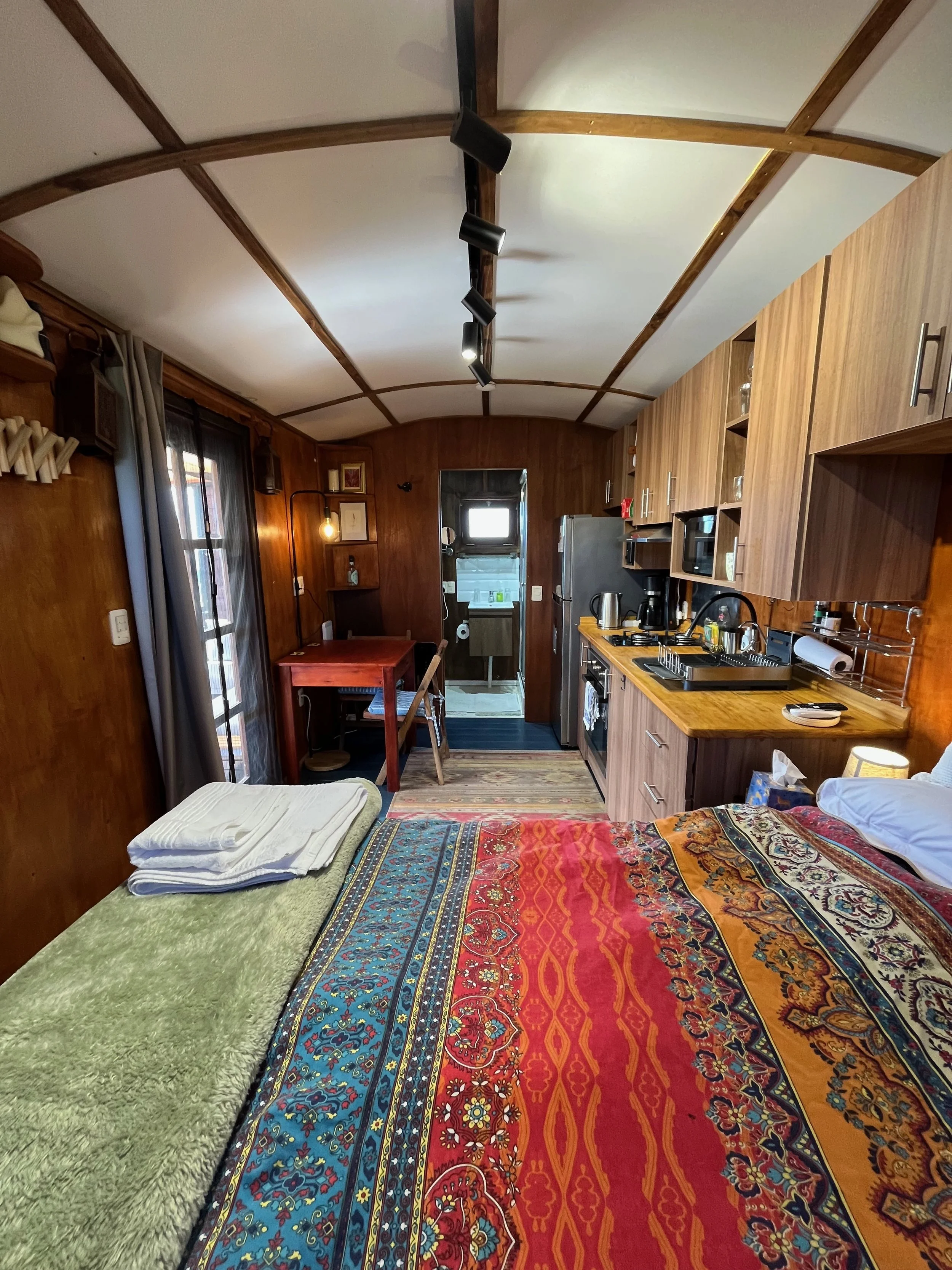 Cozy wooden cabin interior with a bed covered in a colorful, patterned quilt, a small sink and kitchenette, and a window with curtains, giving a warm, rustic feel.