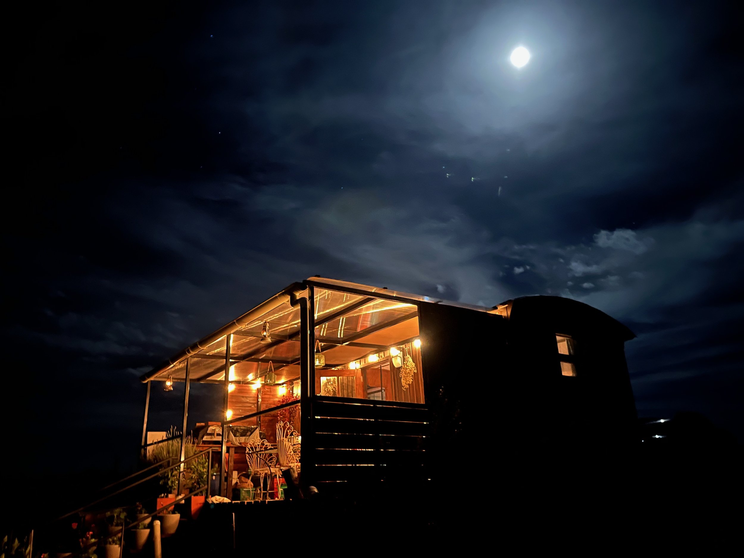A house on a hill illuminated at night with the full moon and clouds in the sky.