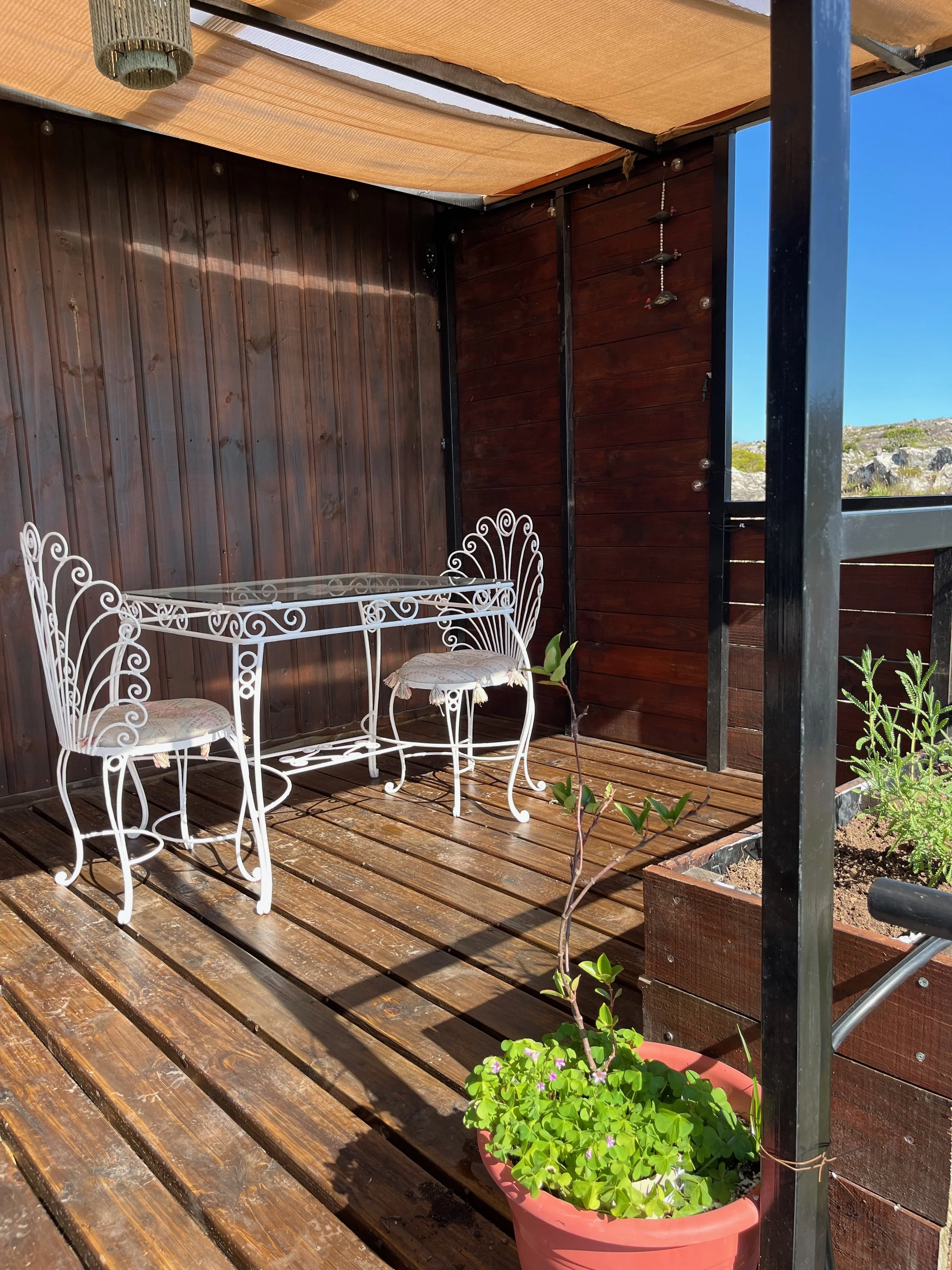 A small outdoor patio with a wooden floor, enclosed by dark brown wooden walls and a beige canopy. There is a white wrought iron table and two matching chairs with upholstered cushions, a pink potted plant with green leaves, and a small garden bed wi