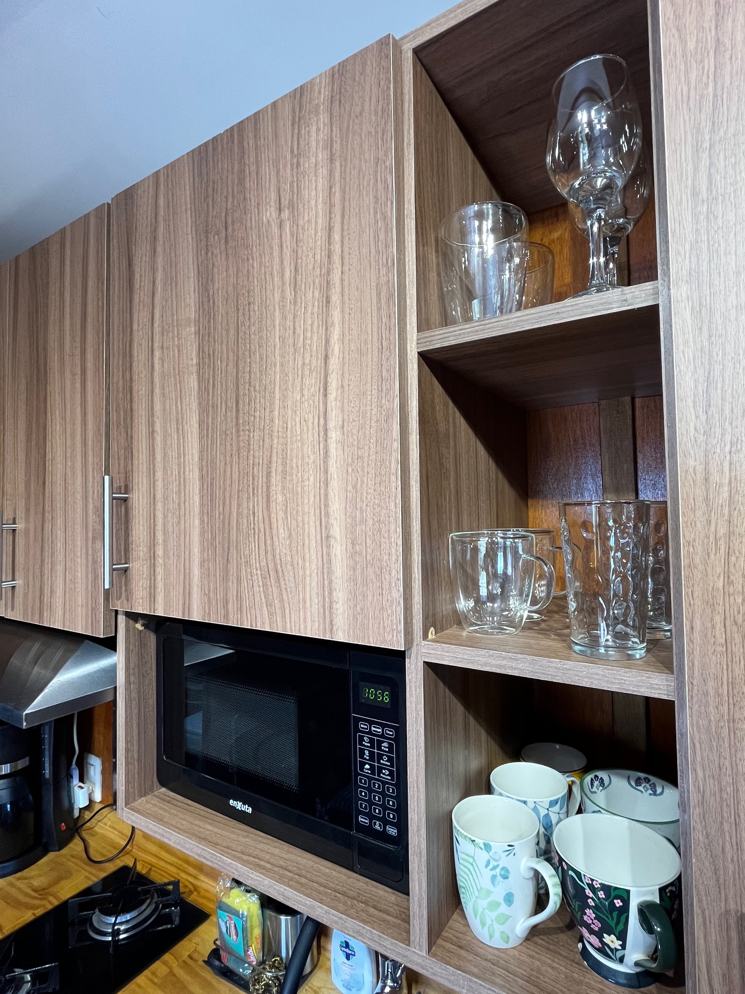 Kitchen cabinet with open shelves displaying glassware and mugs, microwave oven embedded in wood cabinet, and a portion of a stove and coffee maker on the counter.
