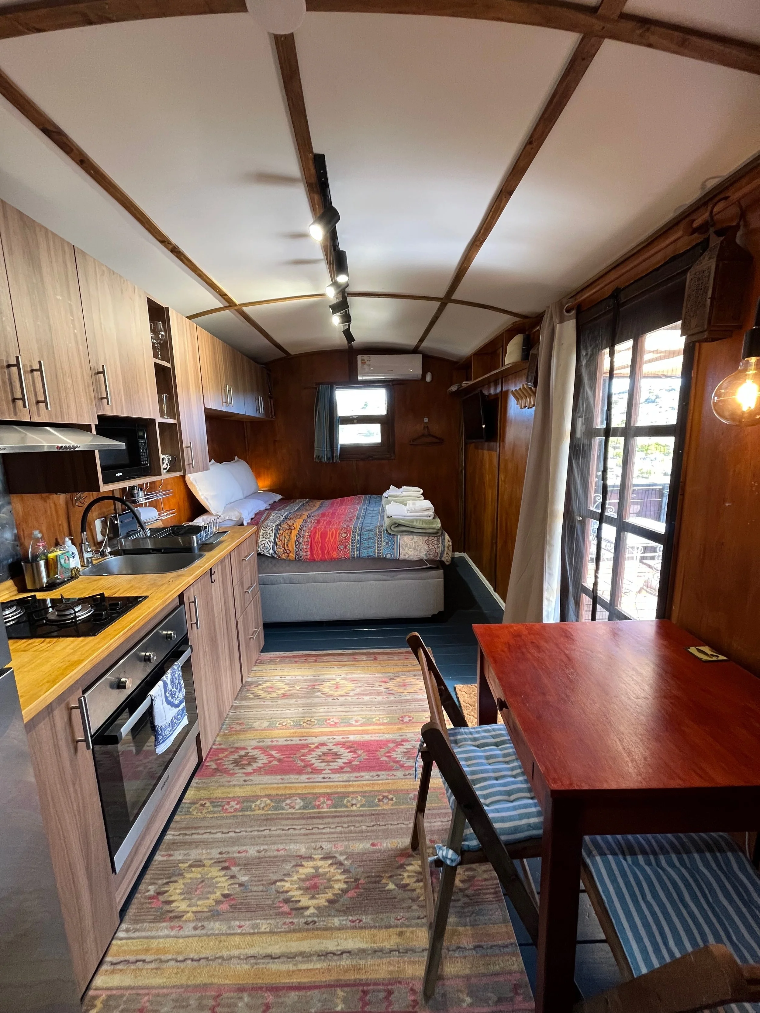 A cozy studio apartment with wooden walls, a kitchenette on the left, a bed in the center, a small dining table with two chairs on the right, a window with curtains, and a sliding glass door leading outside.