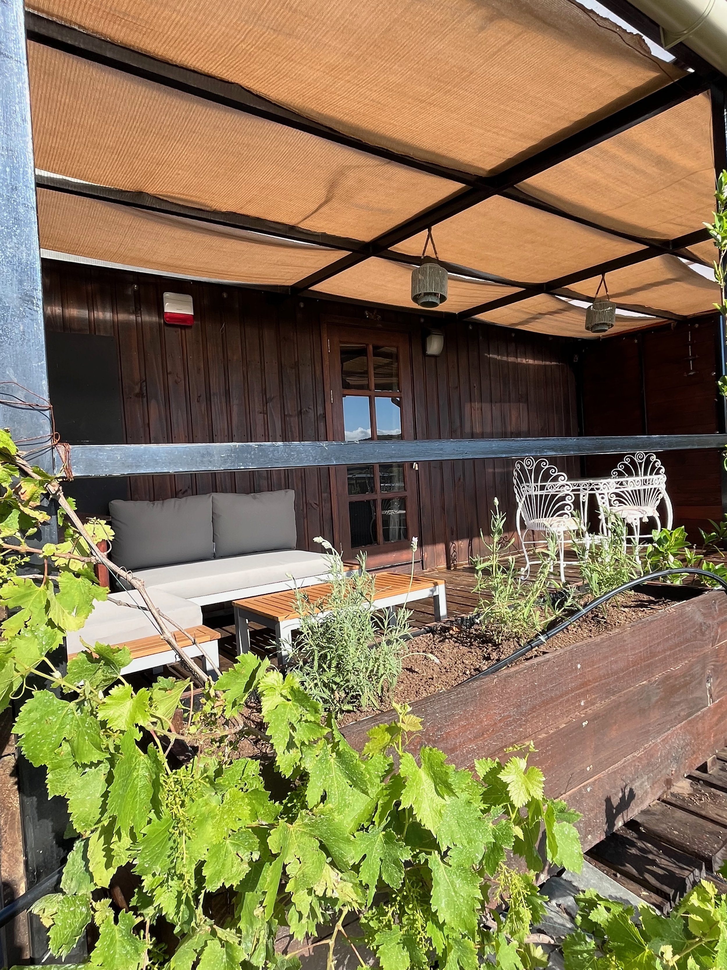 A cozy outdoor balcony with wooden flooring and a covered roof. It features a gray outdoor sofa with cushions, a small wooden coffee table, and ornate white metal chairs. There are plants with green leaves in a long planter box in the foreground.