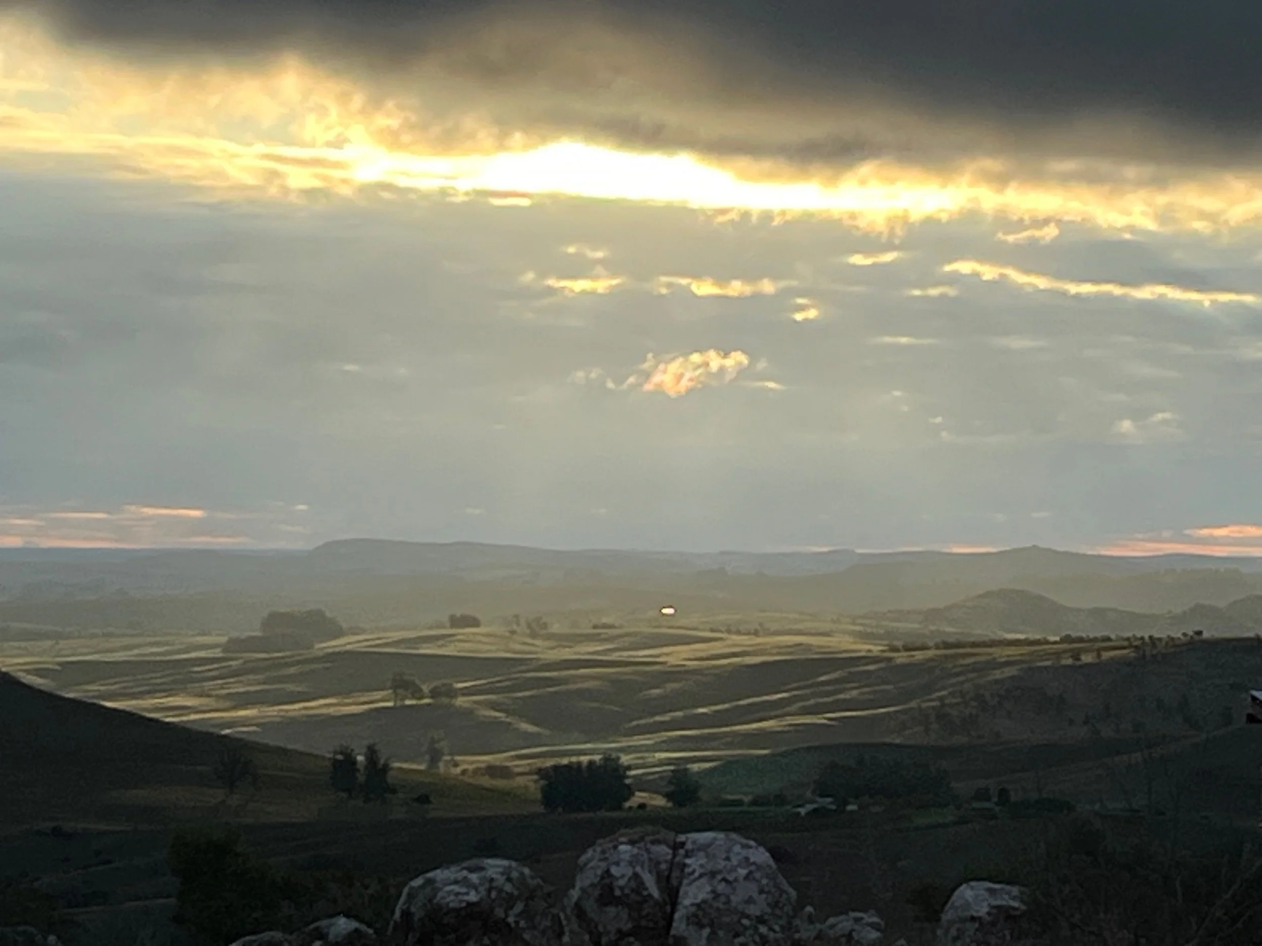 Scenic landscape with rolling green hills and scattered trees under a cloudy sky at sunset.