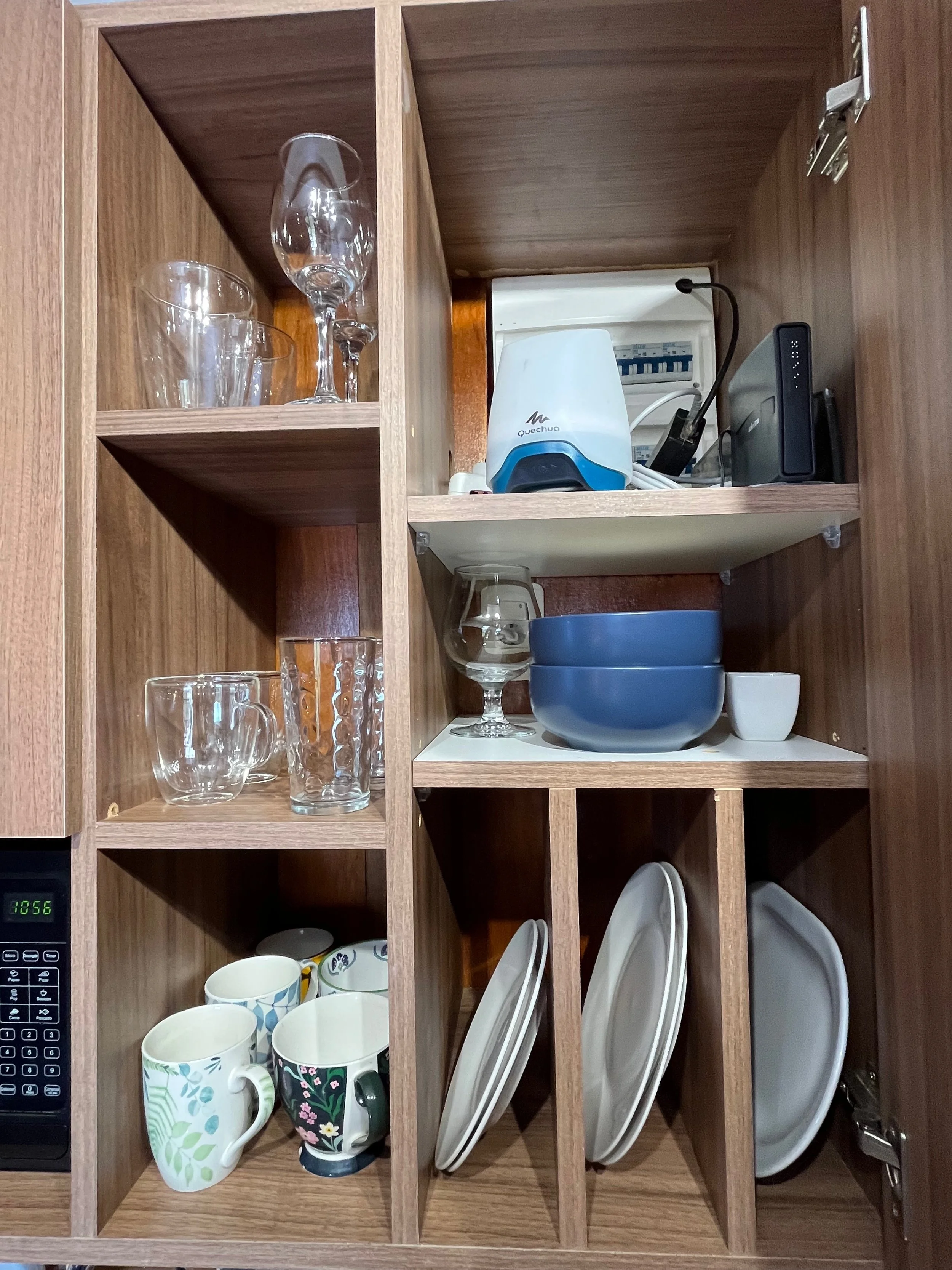 Open kitchen cabinet door revealing dishes, glasses, bowls, cups, and miscellaneous kitchen items inside.