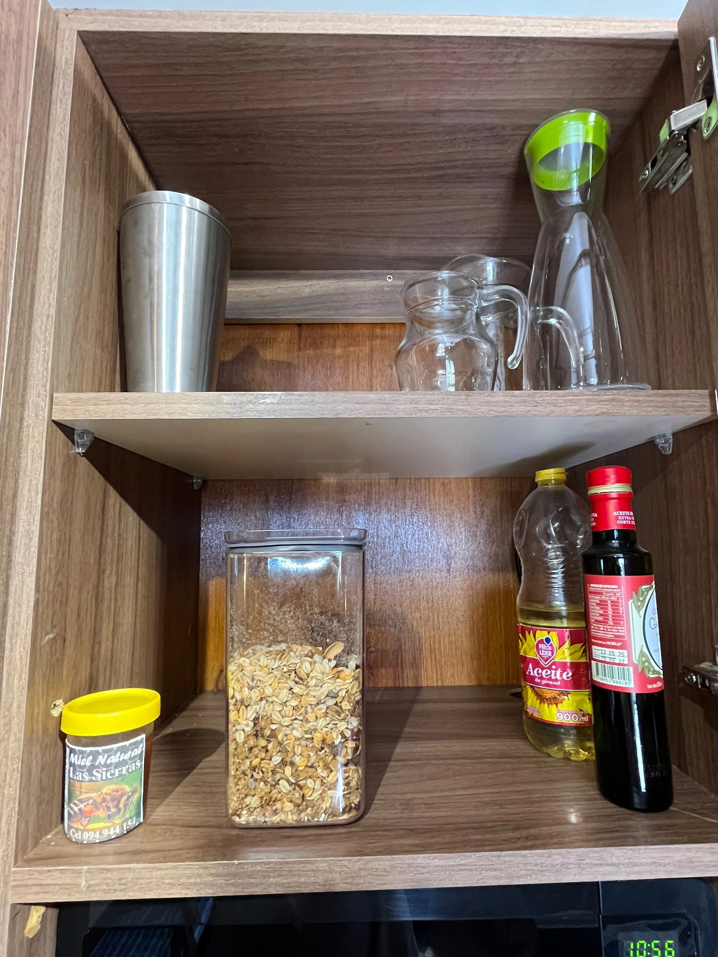 Open wooden cabinet with two shelves containing various kitchen items: a stainless steel tumbler, glass pitcher and cup, a glass pitcher with a green lid, a bottle of sunflower oil, a bottle of soy sauce, a small container of honey, and a glass jar o