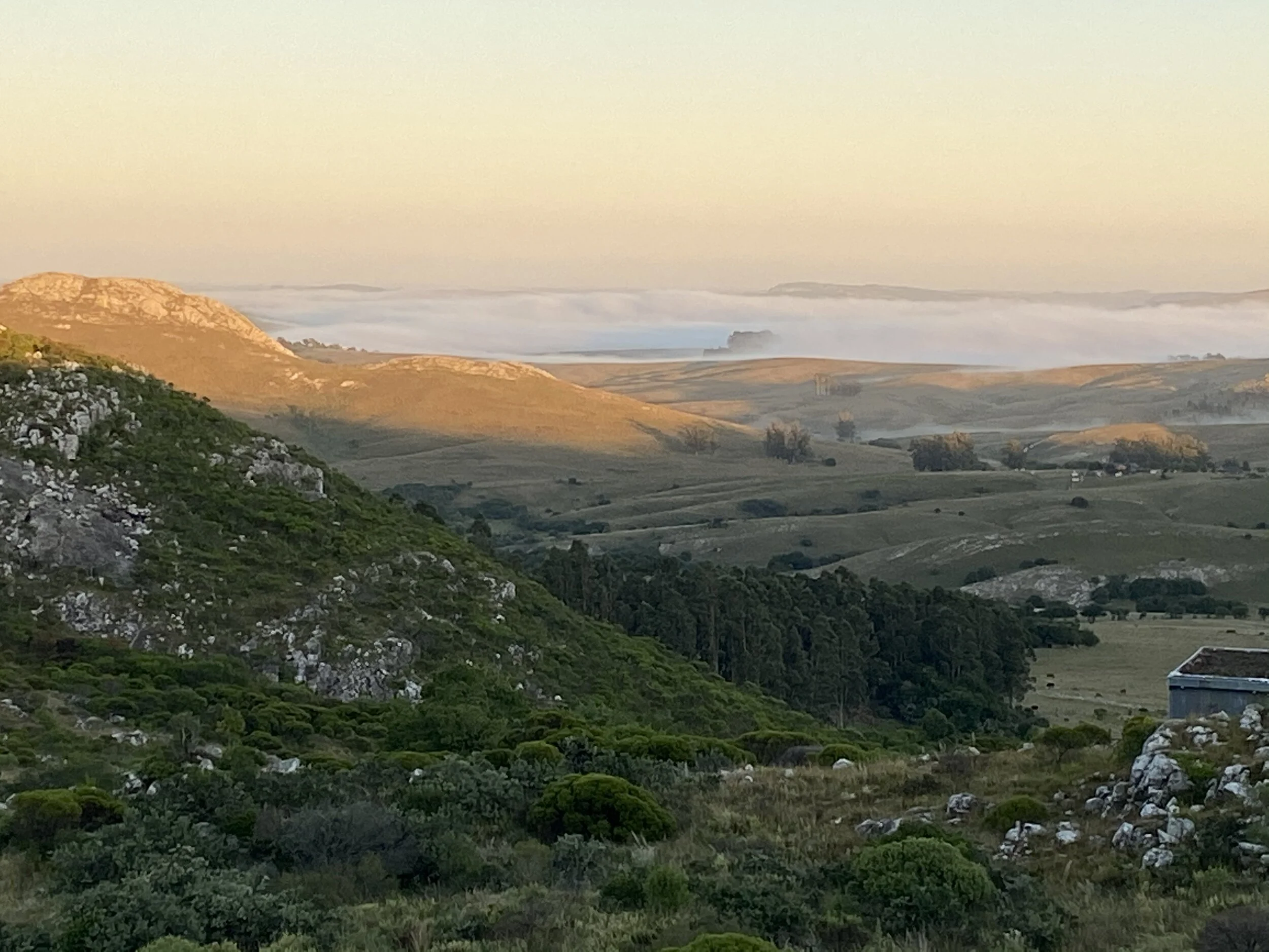 Scenic view of rolling hills and mountains with green vegetation, rocks, and a small building, under a sky with clouds and fog in the distance during sunrise or sunset.