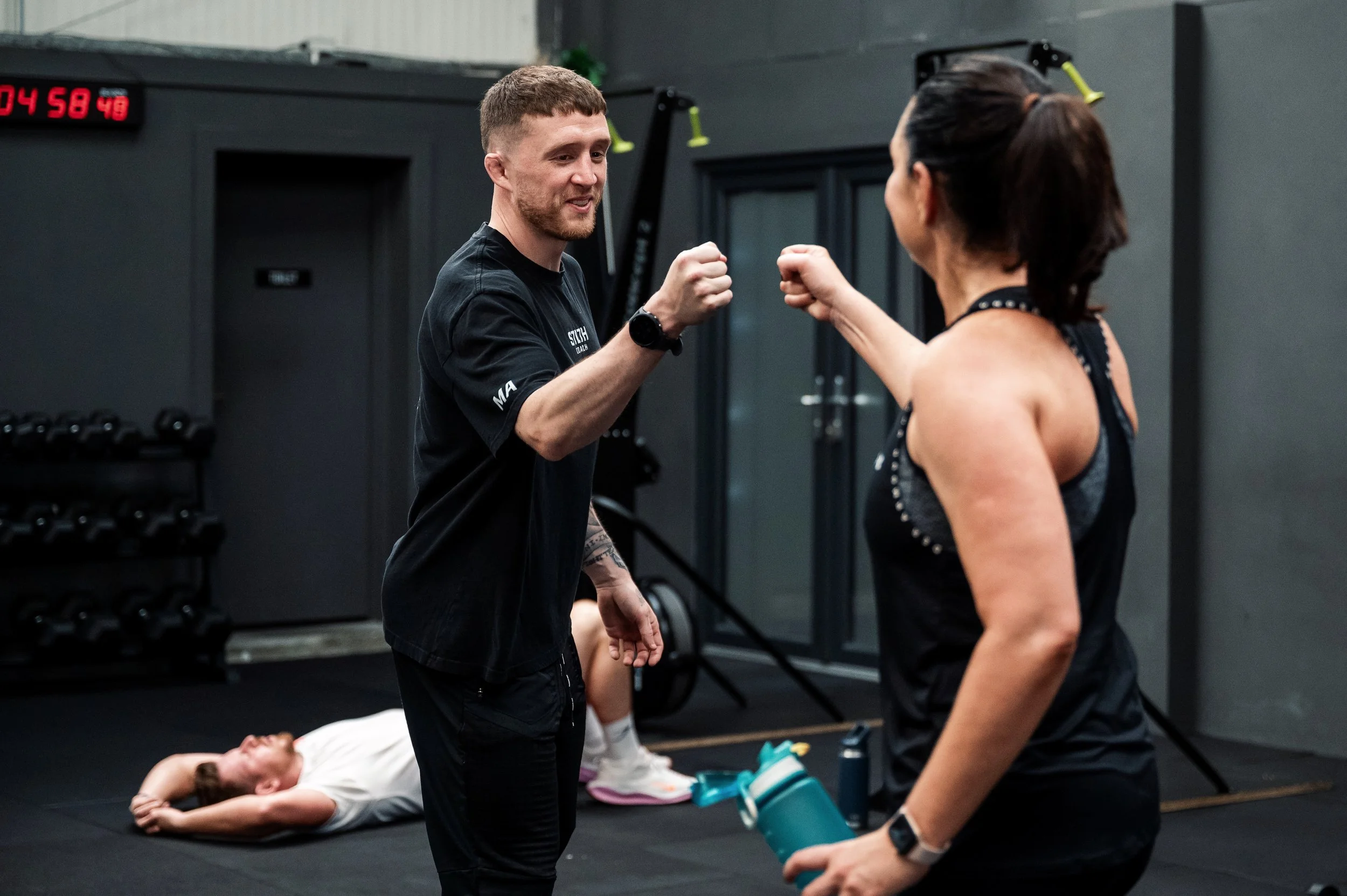 A man and woman in a gym, greeting each other with a fist bump, while a woman lies on the floor resting after a workout. The gym has black walls, weights, and a digital clock in the background.