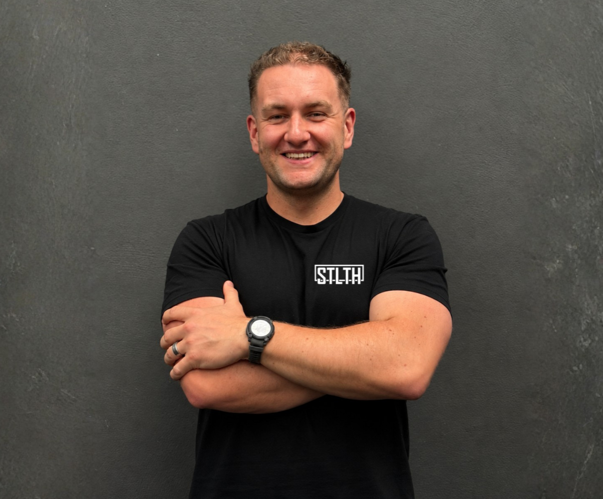 Ethan Savage: a smiling man with short, curly hair standing against a dark grey wall. He is wearing a black T-shirt with a logo that says 'SLTTH' on the front, a black wristwatch, and a wedding ring. His arms are crossed.
