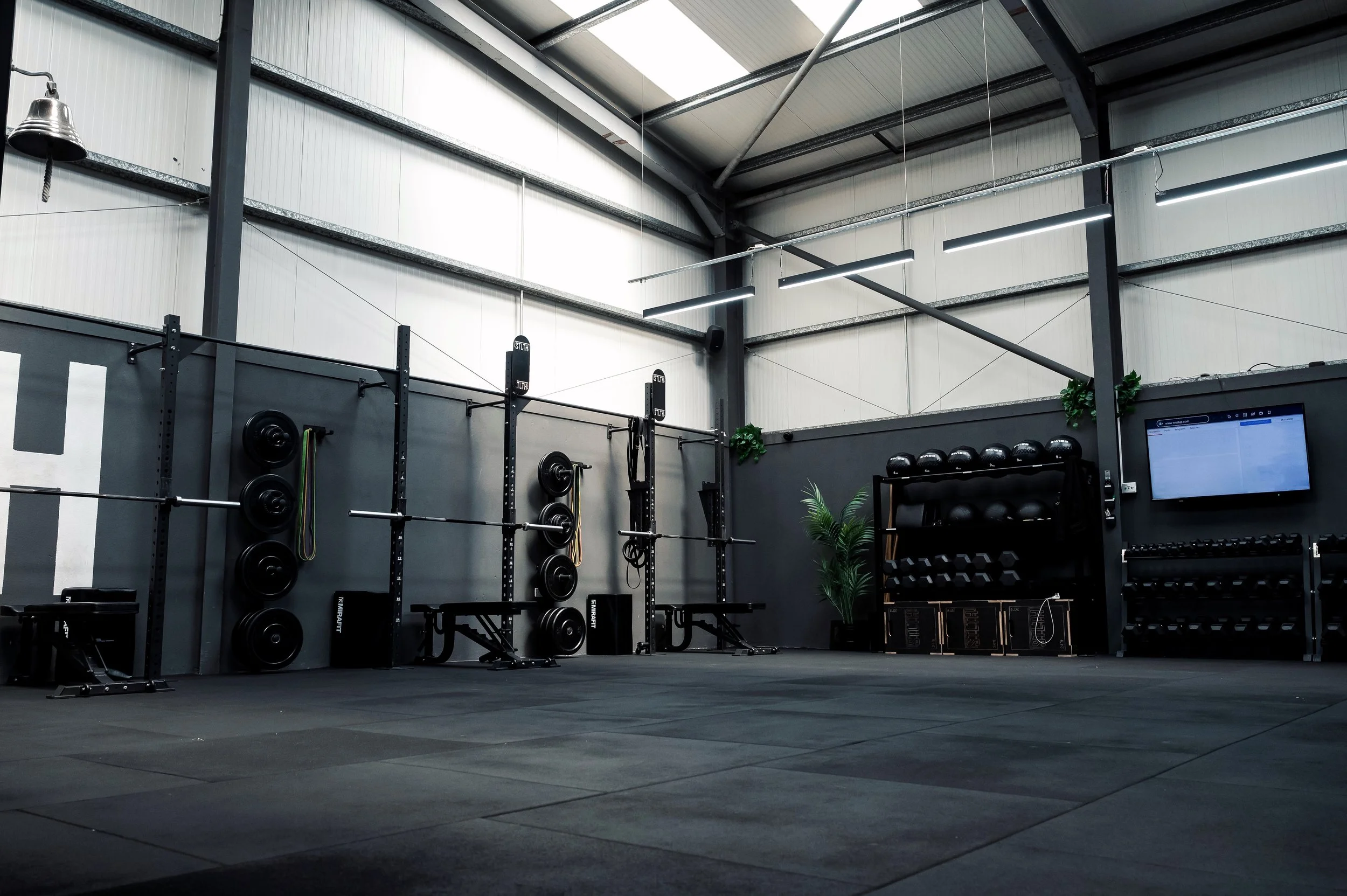 Interior of a modern gym with weightlifting equipment, black rubber flooring, and bright overhead lighting.