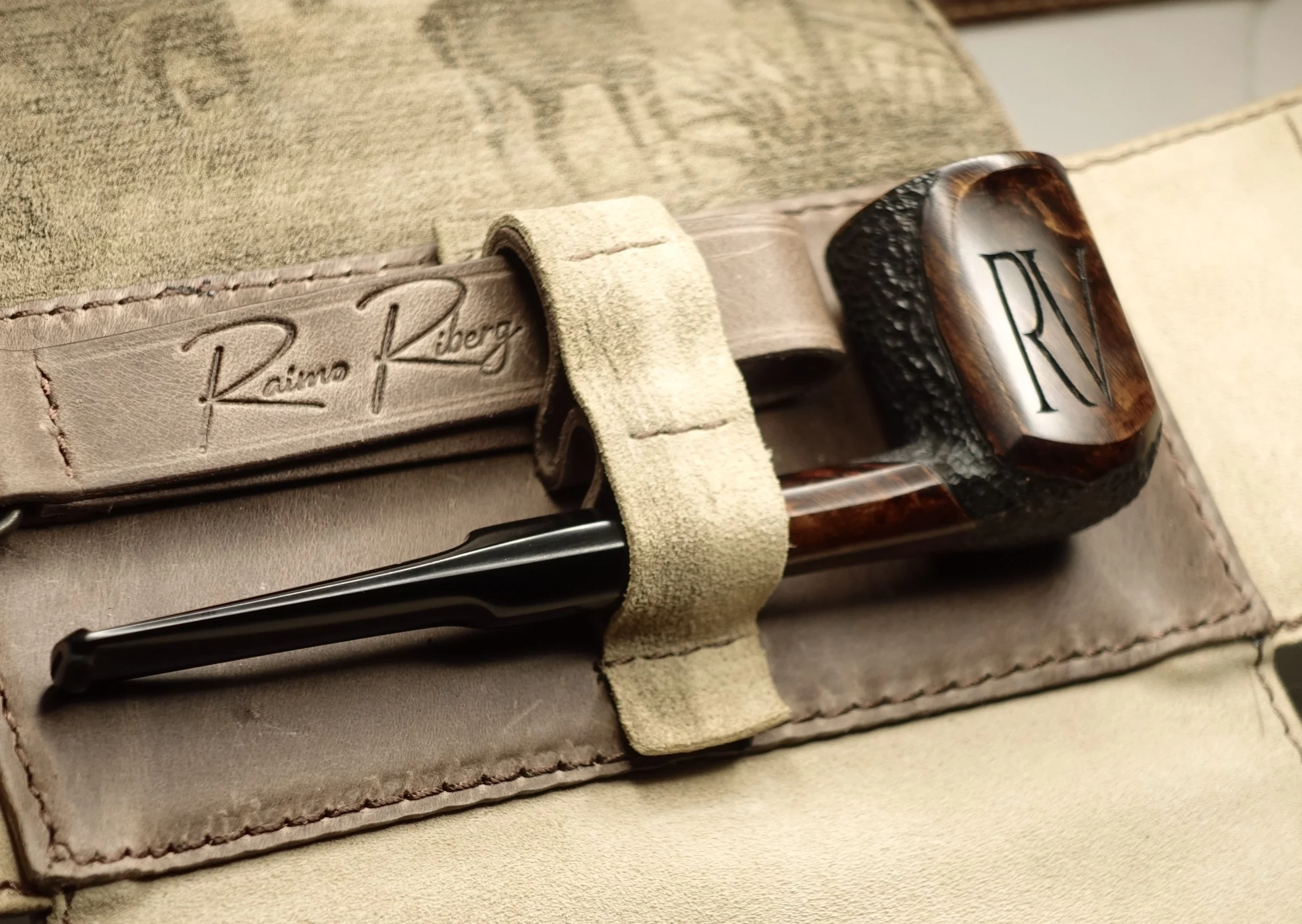 Close-up of a wooden pipe with carved initials "RV," secured in a leather holder with "Raimo Riberg" branding.