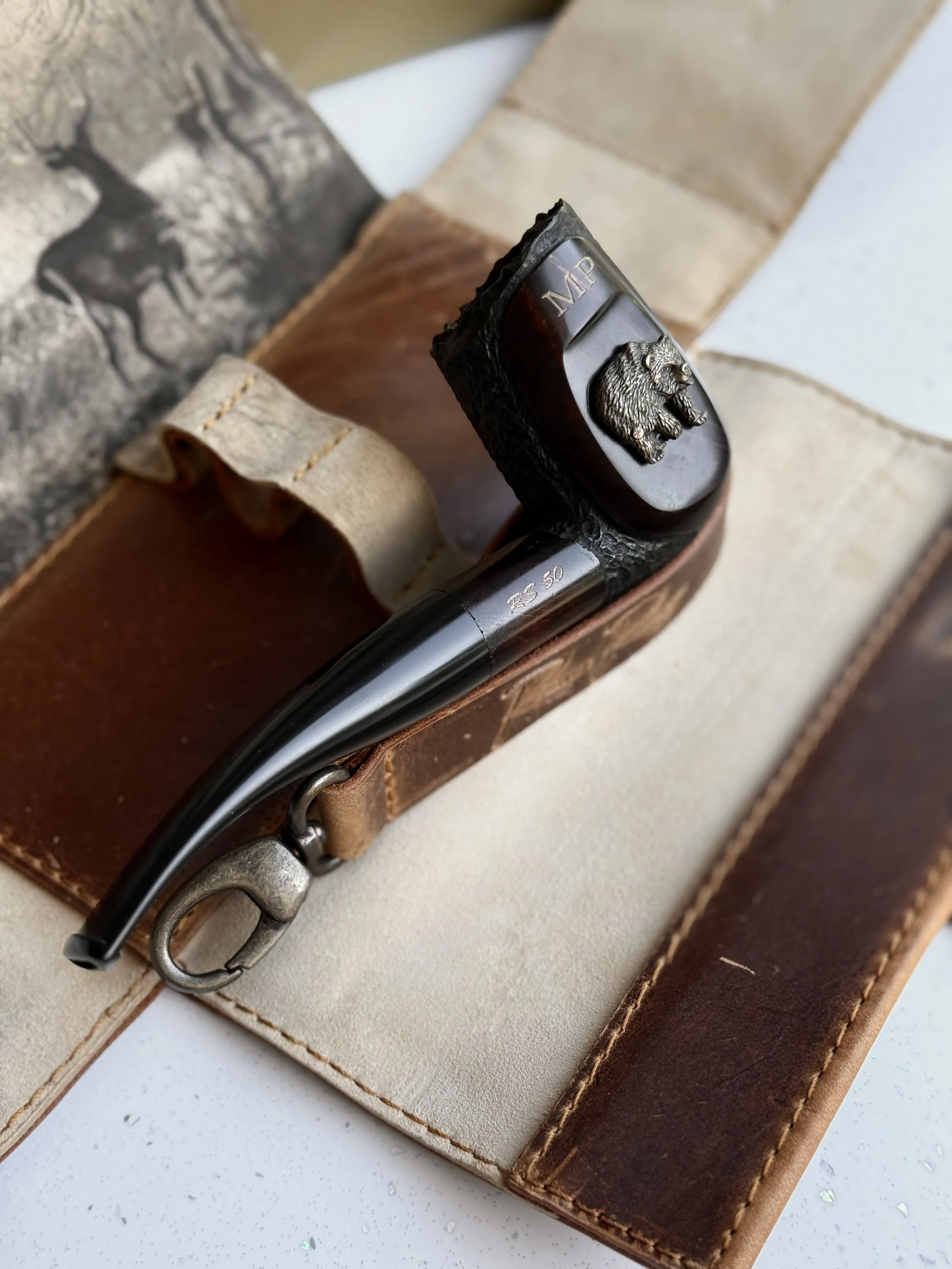 Luxury handmade briar tobacco pipe from the Riberg MP Wildlife Series, crafted in Estonia and inspired by forests, wildlife, and nature. a leather pipe with a deer head emblem, a leather belt, and a leather bag.