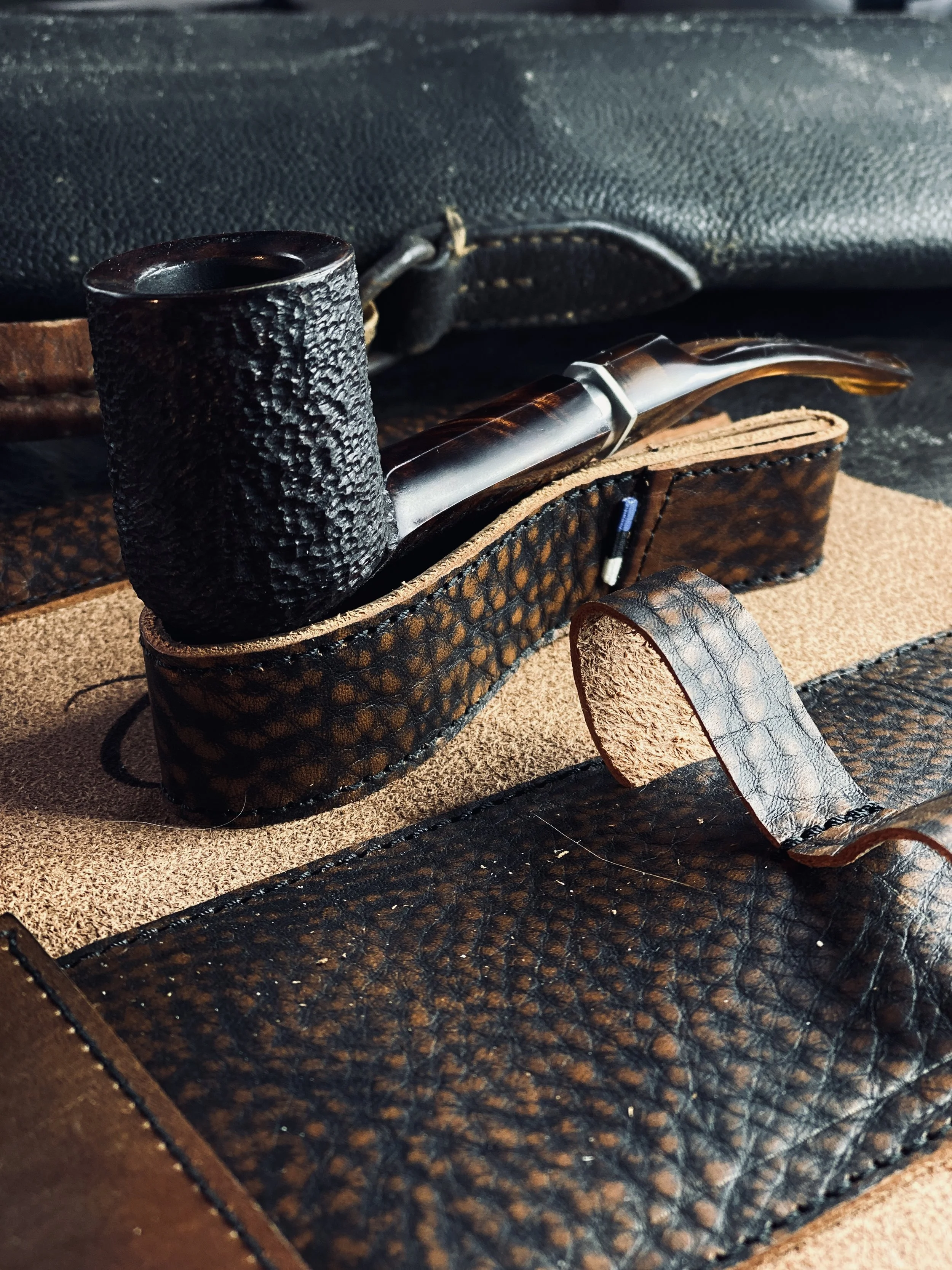 Close-up of a textured leather pipe with a black and brown design, against a background of various leather items. The pipe has a rustic, handcrafted appearance.