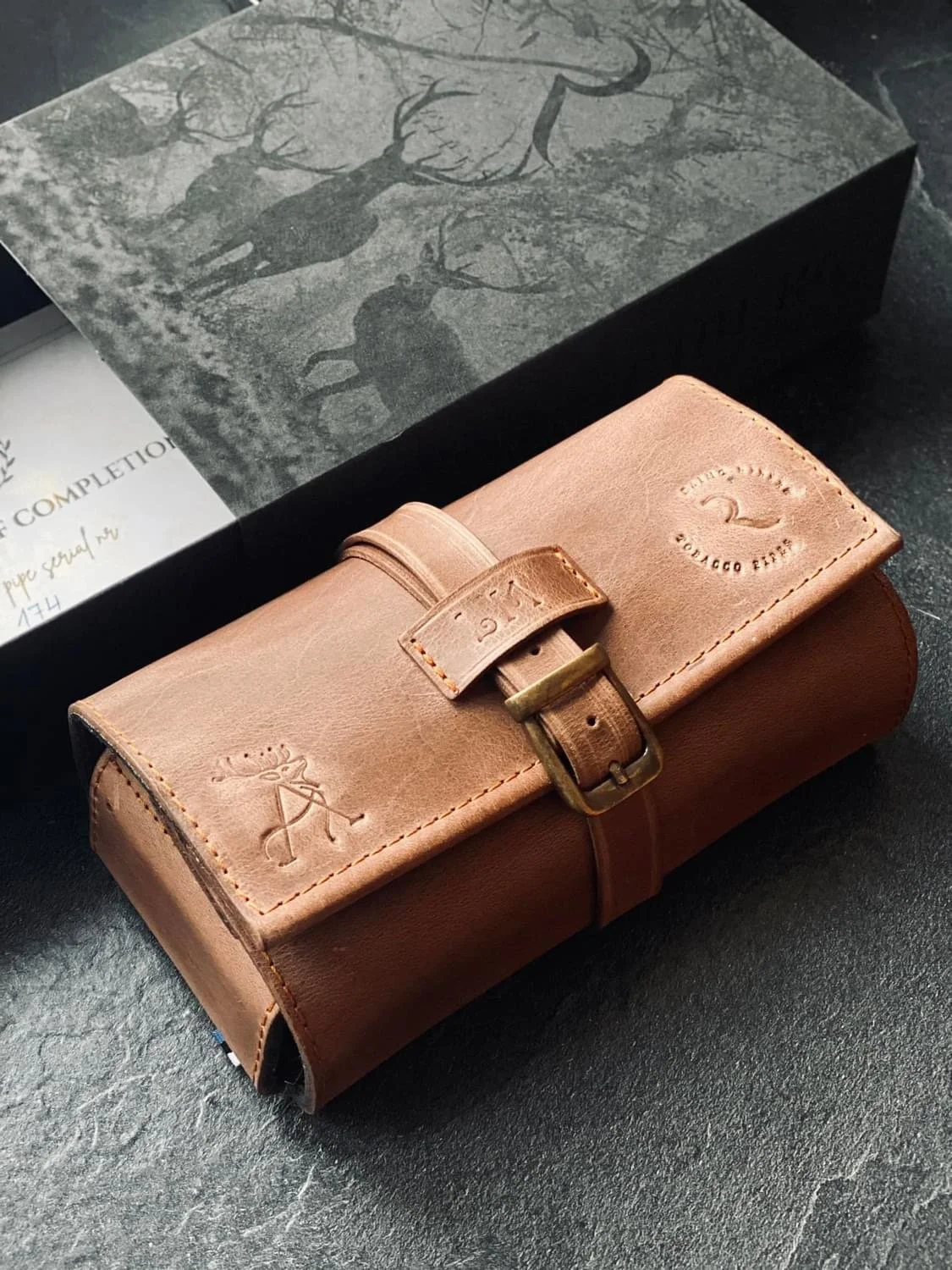 Leather watch roll with buckle, embossed with deer and logo, on dark surface in front of a black box featuring deer silhouettes.