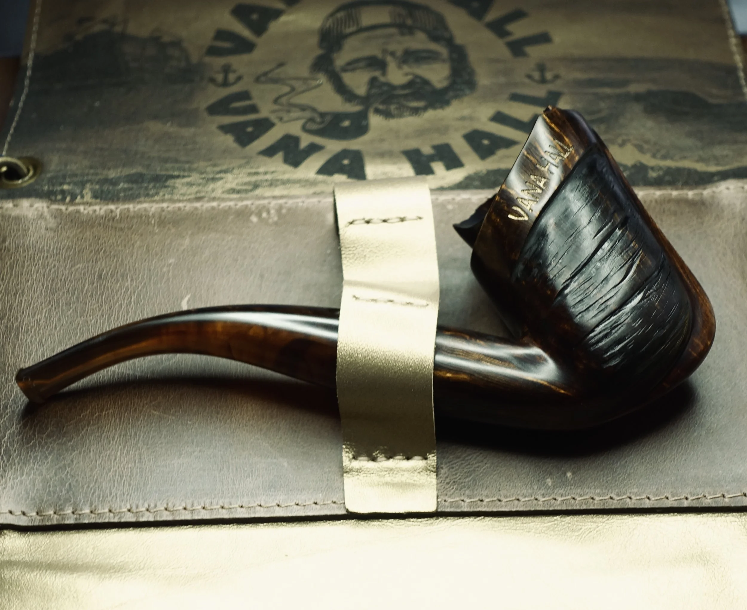 A smoking pipe with a dark brown, textured bowl and a curved stem resting on a leather surface, with a vintage-style paper bag in the background.