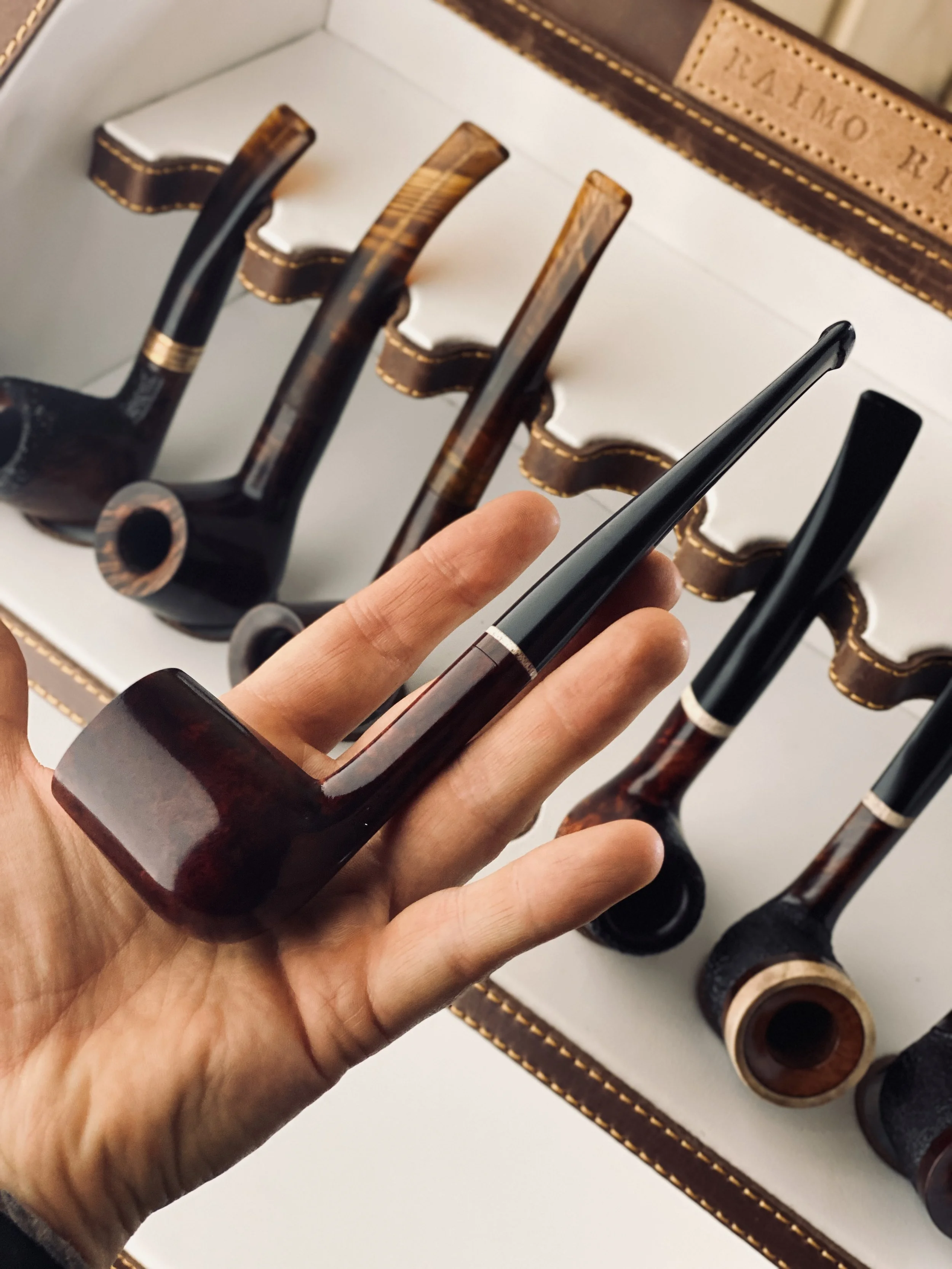 Hand holding a wooden smoking pipe in front of a display of similar pipes on a white and brown case.