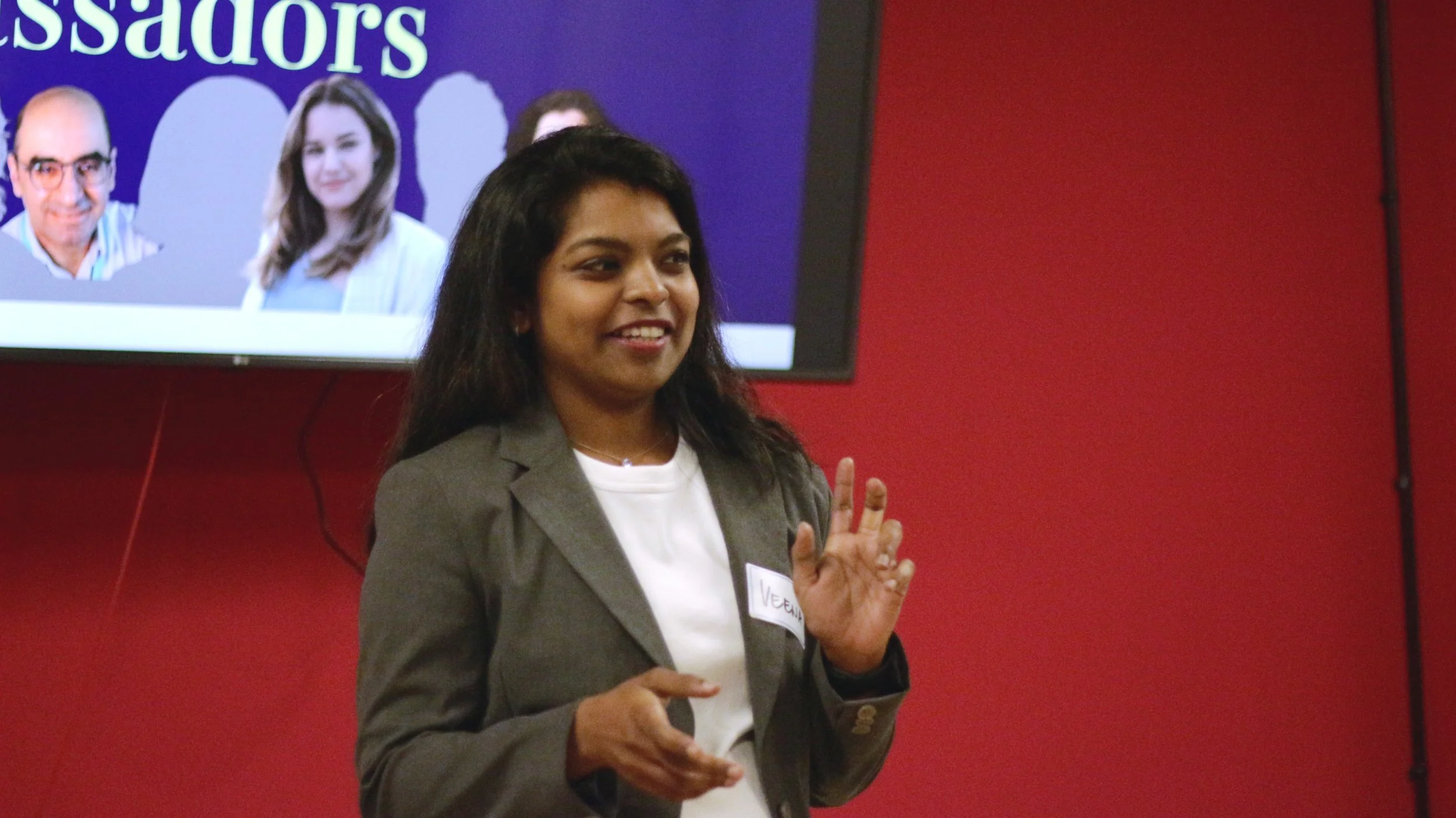 A woman speaking at a presentation or event, with a red wall and a screen displaying headshots behind her.