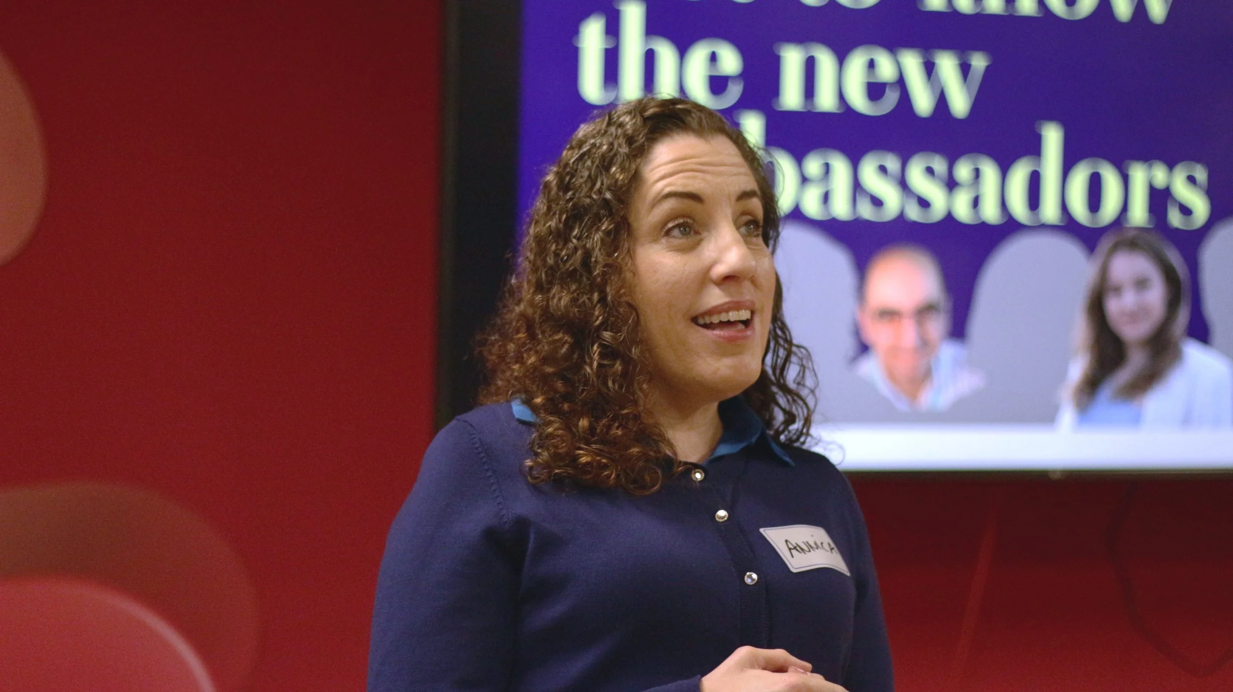 A woman with curly brown hair and a name tag labeled "Annie" is speaking in front of a screen that reads "the new ambassadors" and shows pictures of three people.