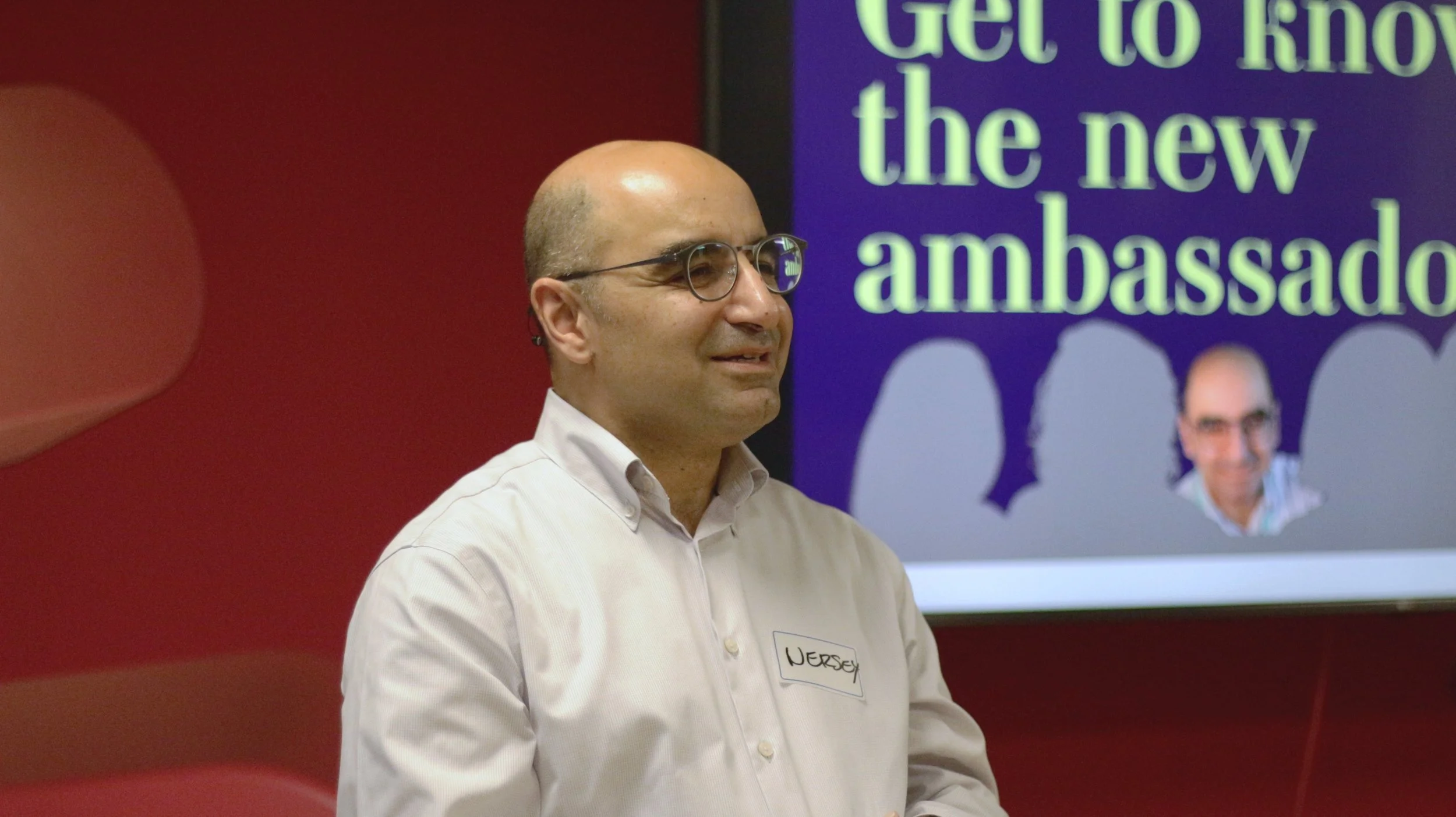 A man with glasses and a white shirt standing in front of a screen that reads 'Get to know the new ambassador.'