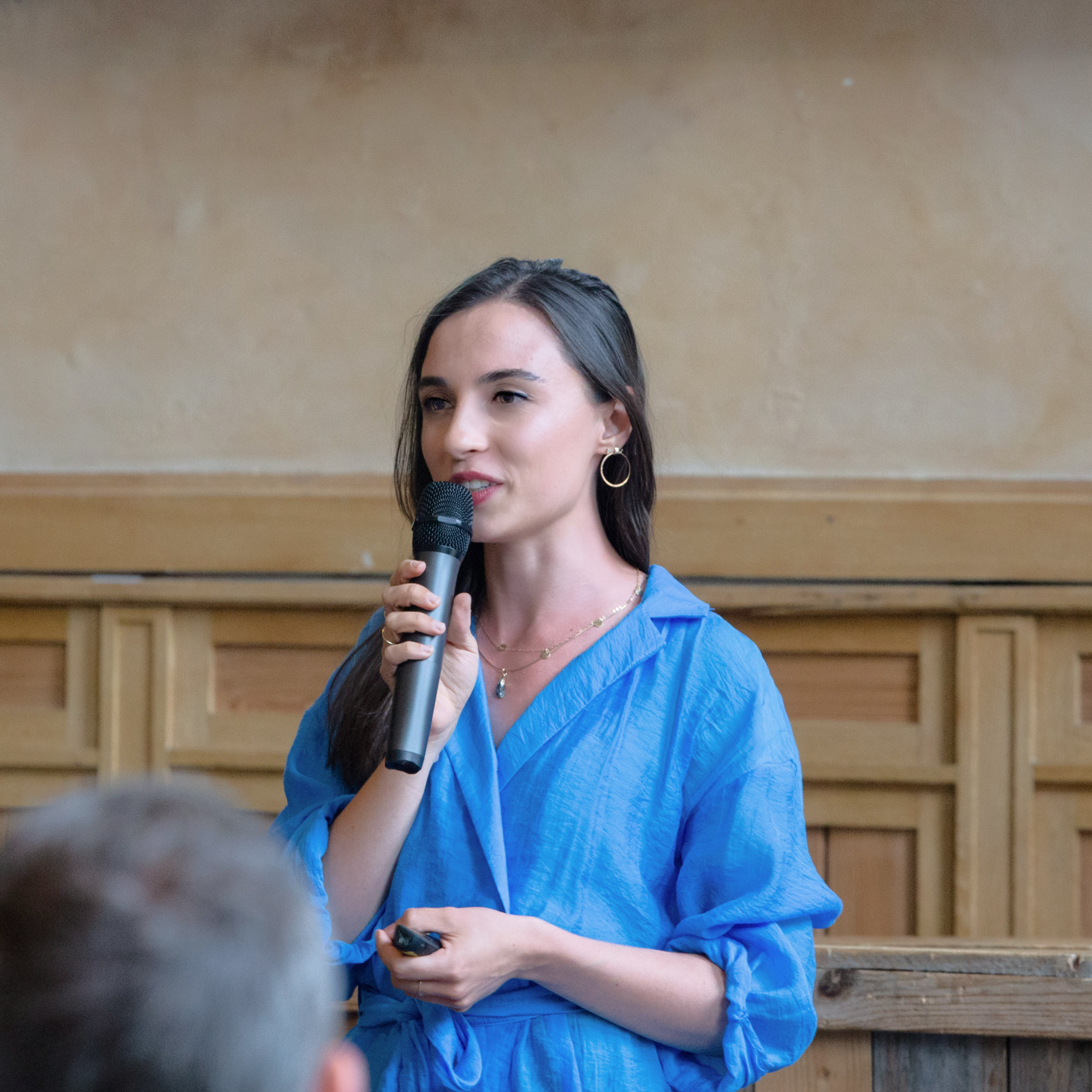 A woman with long dark hair, wearing a blue shirt, speaking into a microphone during a presentation in a room with wooden paneling.