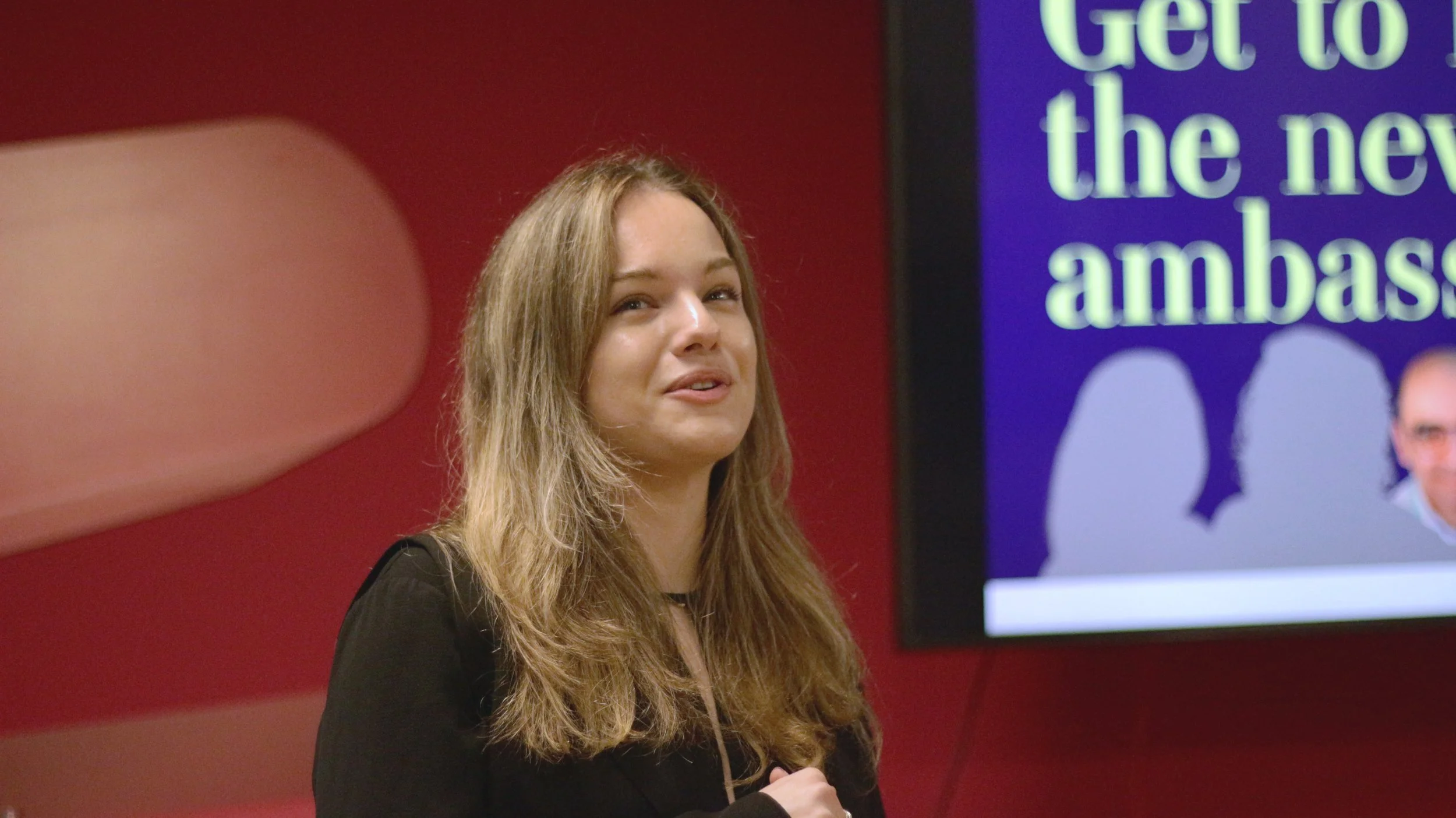 A woman with long wavy light brown hair and a black top standing in front of a red wall, with a large digital screen displaying partially visible text and pictures of people.
