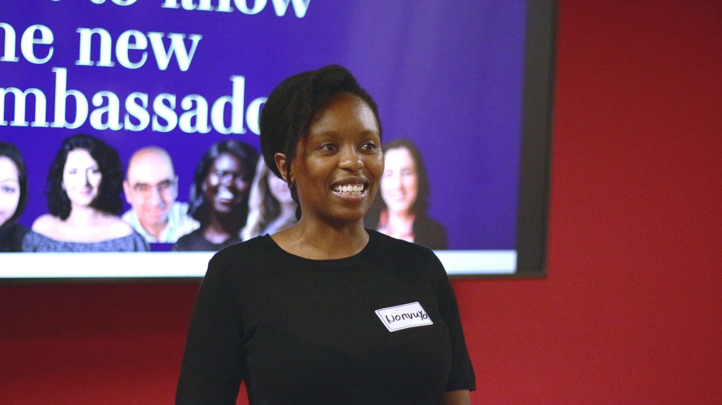 A woman smiling, wearing a black shirt with a name tag, standing in front of a screen displaying a diverse group of people and the text 'the new ambassador'.