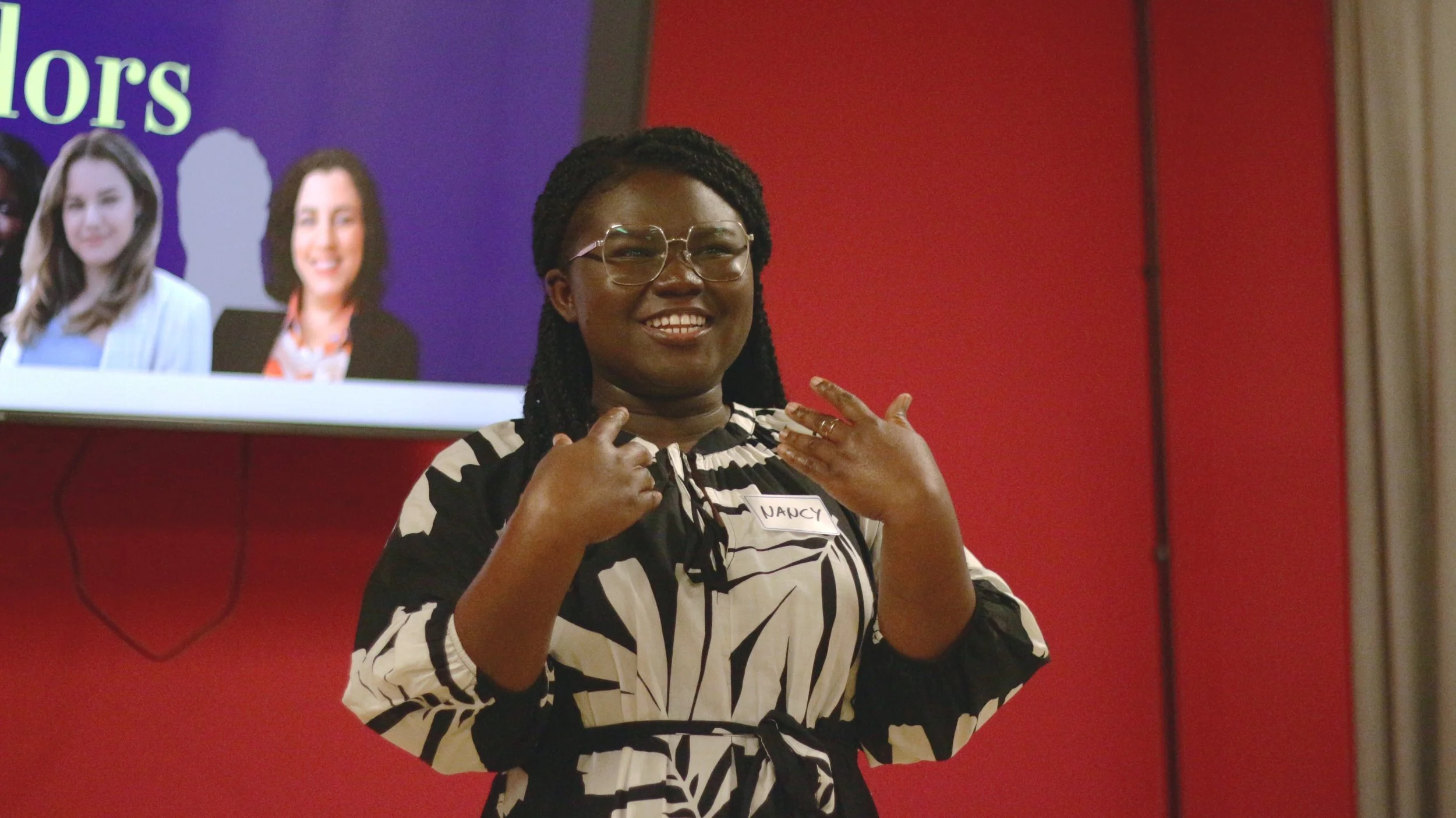 A woman smiling, wearing glasses and a black-and-white patterned dress with a name tag that reads "Nancy," standing in front of a red wall and a screen displaying images of other women.
