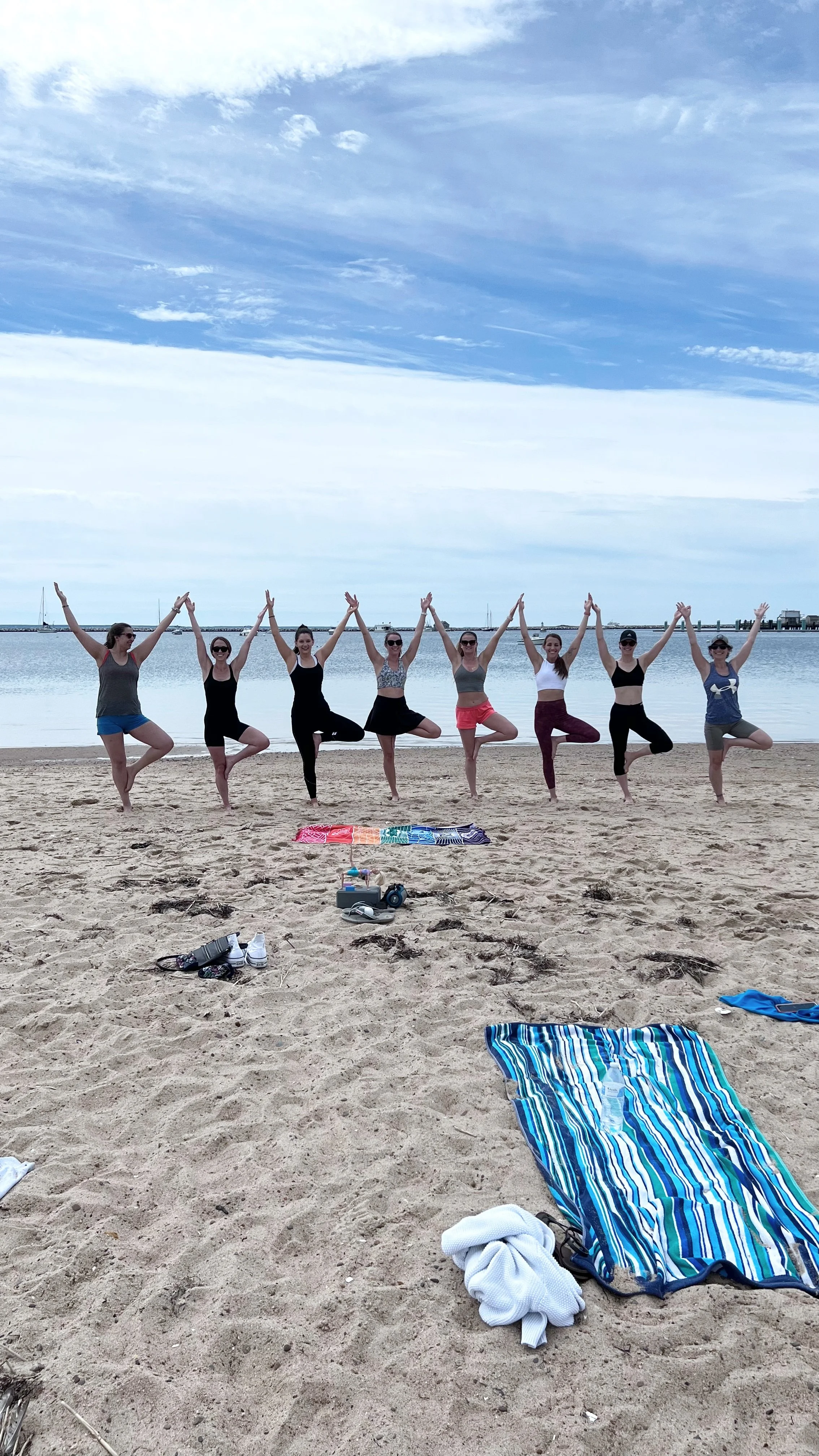 Group of women doing yoga poses on a sandy beach near the ocean, with towels and personal items on the sand.