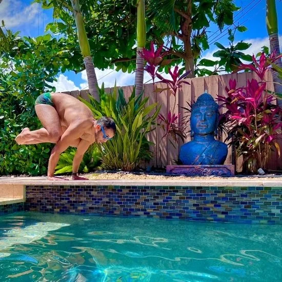 Person wearing swimming goggles and shorts performing a yoga pose by a pool, surrounded by vibrant plants and a blue Buddha statue.