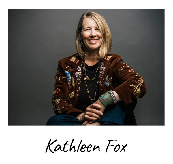 Portrait of Kathleen Fox, a woman with shoulder-length blonde hair, smiling, wearing a brown embroidered jacket, black shirt, and multiple beaded necklaces, sitting against a gray background.