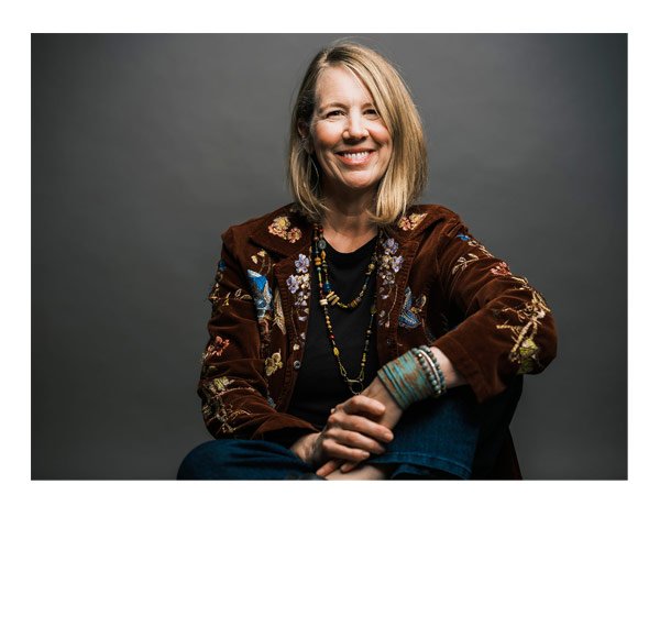 Smiling woman with blonde hair wearing a brown embroidered jacket, layered necklaces, and colorful bracelets, sitting against a gray background.